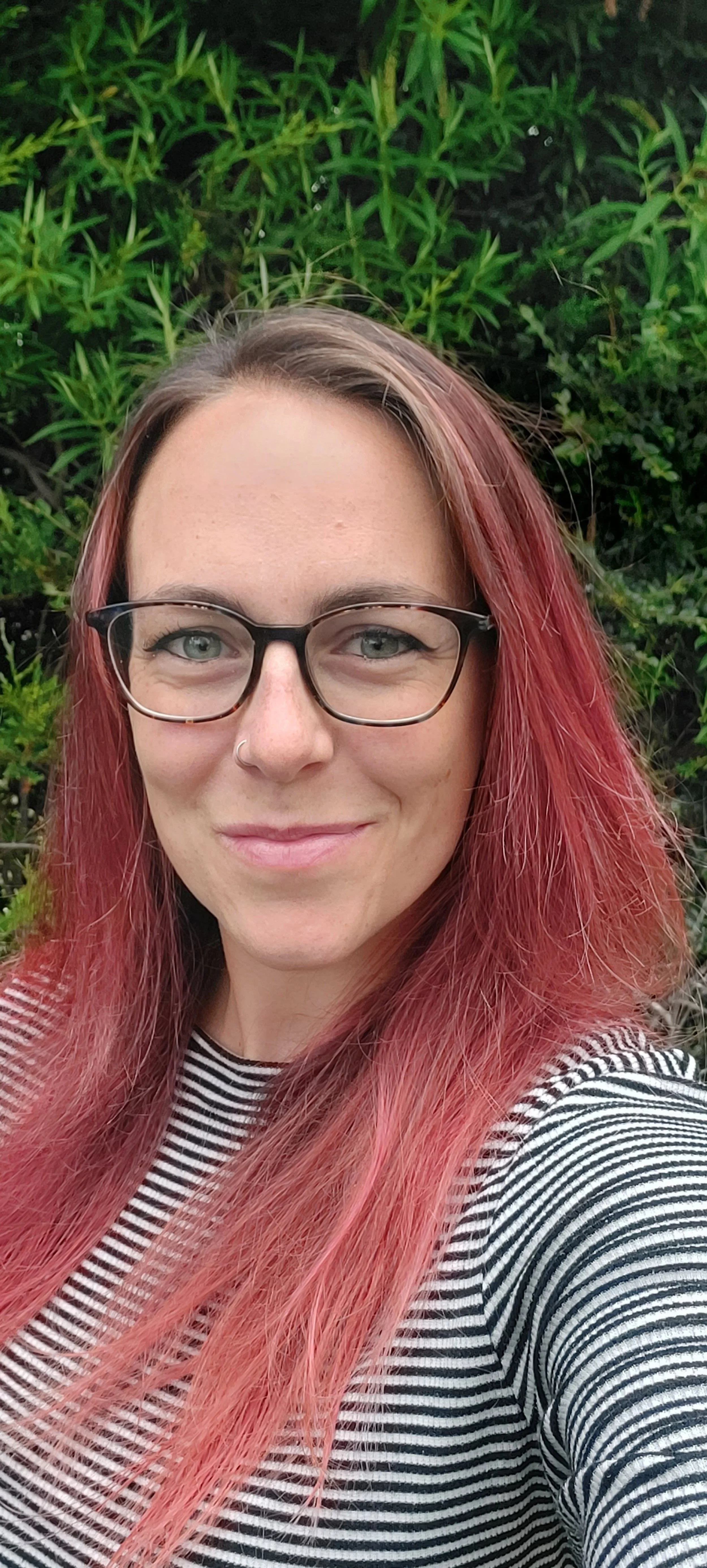 A woman with red hair, glasses, and a nose ring smiling outdoors in front of green foliage.