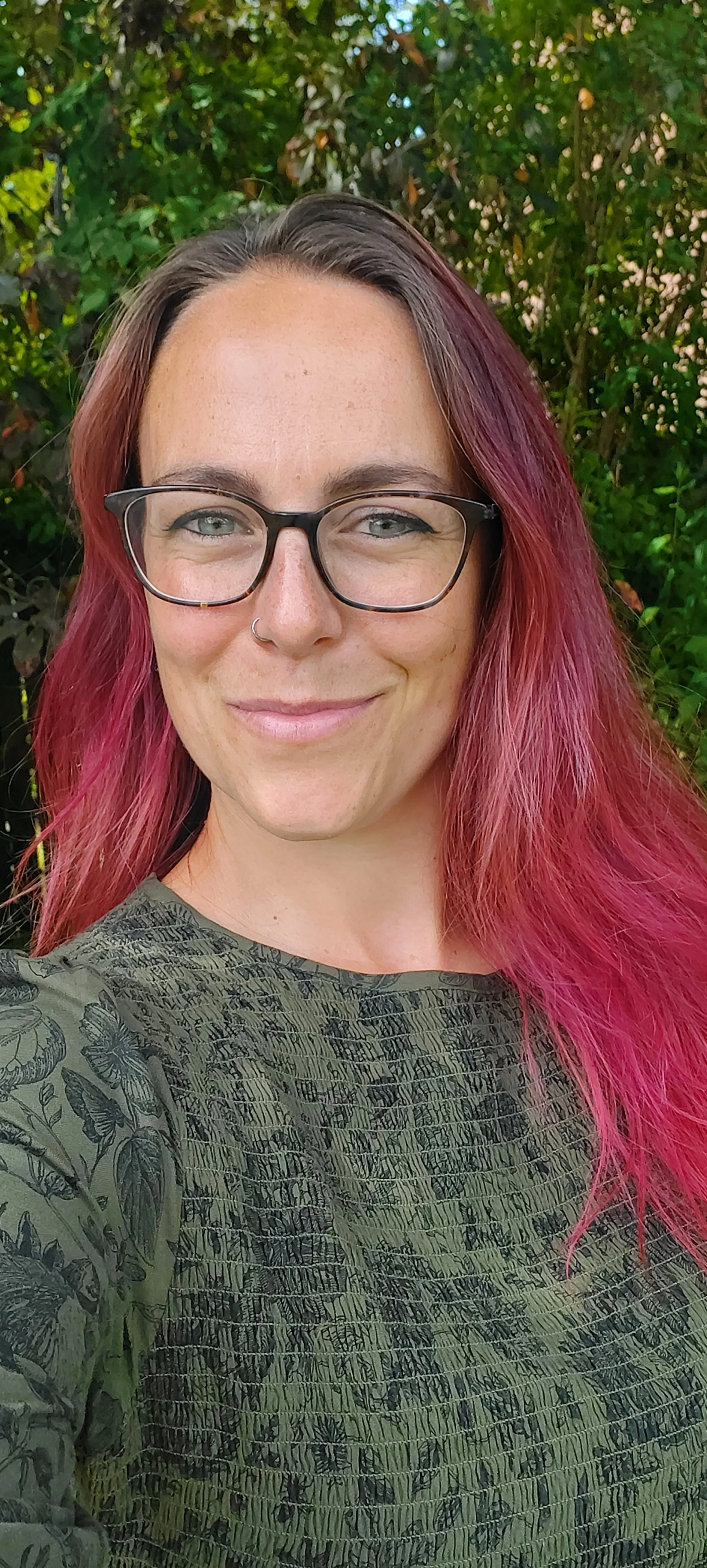 Cassie Hogg, a counsellor with red hair, glasses and a nose ring smiles warmly outside, wearing a green dress.