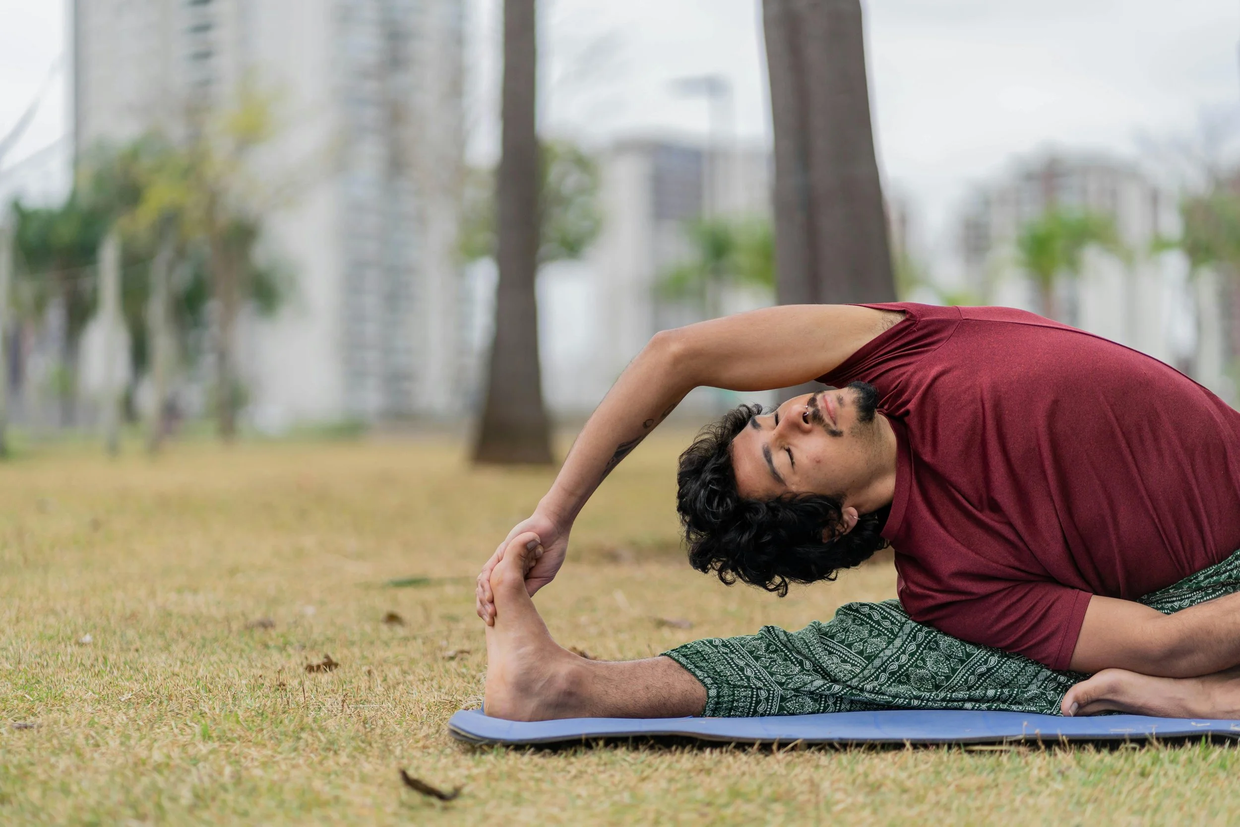 A man doing yoga outdoors on a mat in a park, stretching with one leg extended and holding his foot with both hands.
