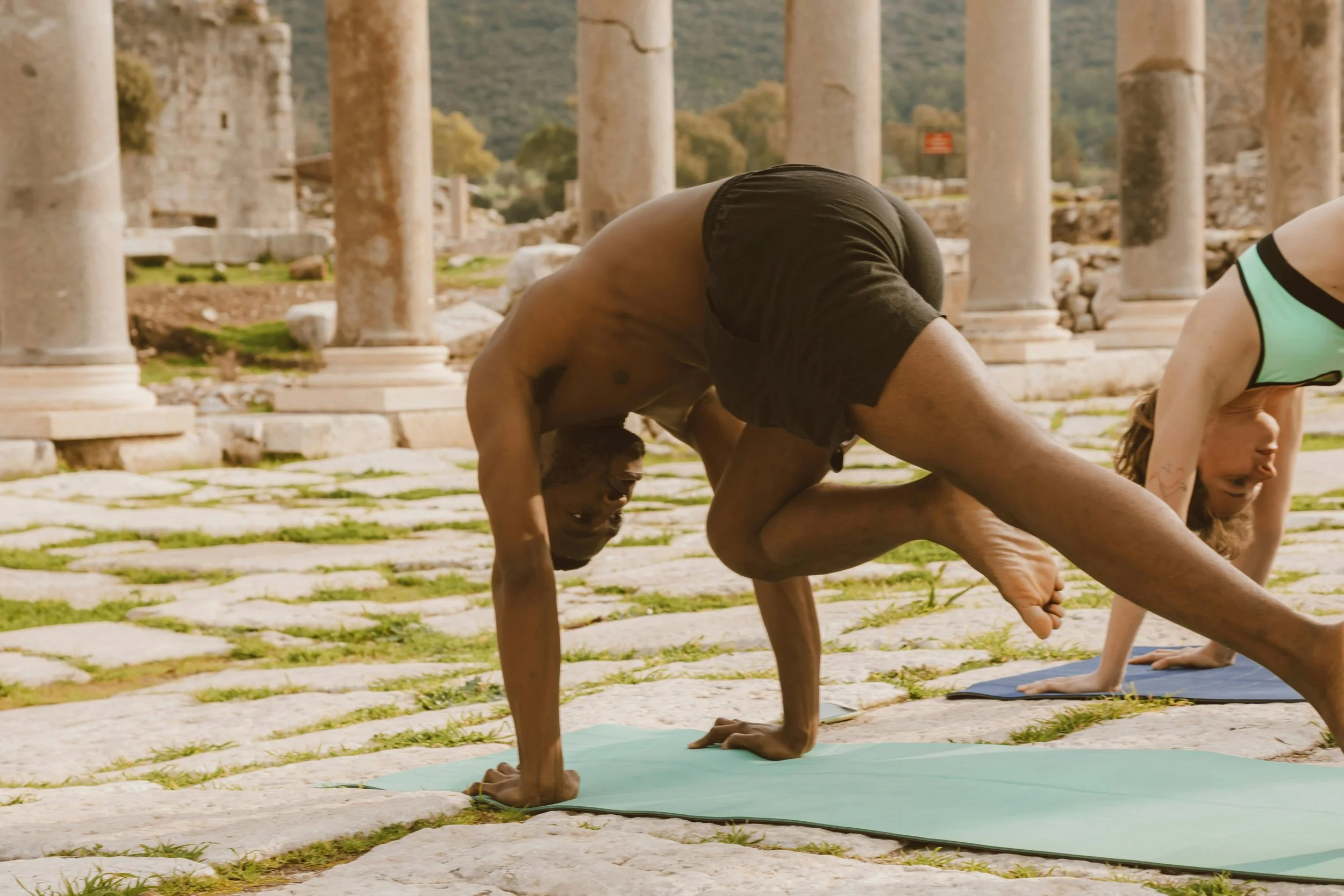 A man doing yoga in a handstand pose outdoors with ancient ruins and stone pillars in the background.