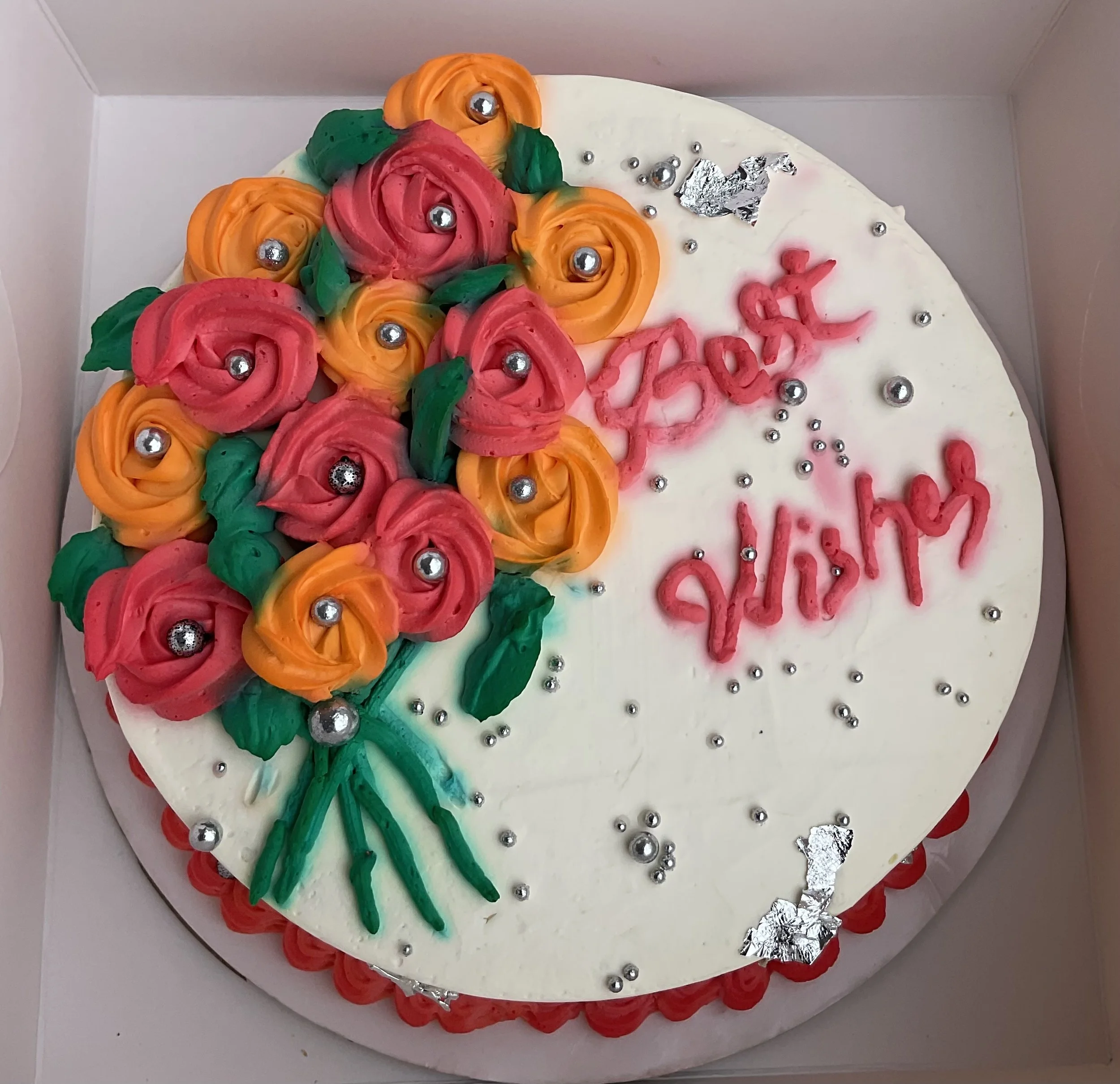 A white cake with pink and orange flowers with green leaves piped between them. Silver foil and edible pearls decorate the cake, which also says 'Best Wishes' in pink frosting.