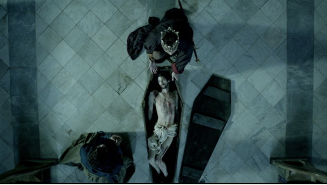 Richard II in the last scene of the Hollow Crown television adaption. The camera looks on the scene from above. Richard's body, bloody and naked save a loin cloth, is in a wooden coffin. Bolingbroke, crowned, sits at his head.