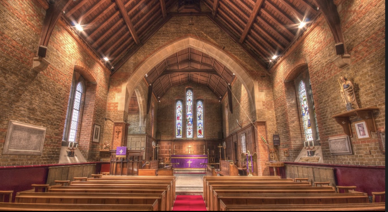 A chapel like space with light brown exposed bricks, a medium brown wooden ceiling, light wood pews and an altar with a purple cloth on it. Behind the altar is 3 stained glass windows which are long and thin.