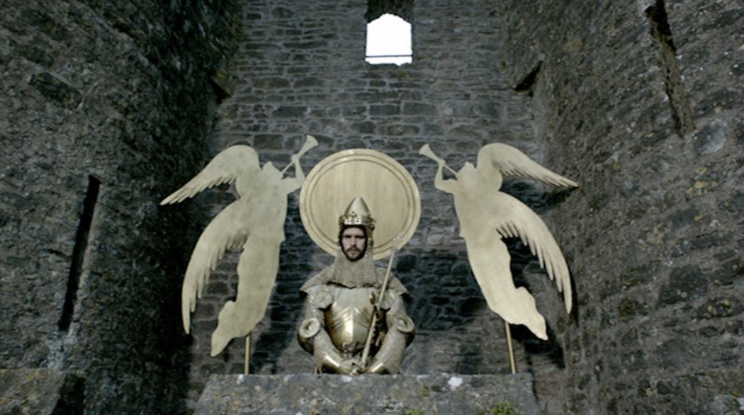 Richard sits at Flint Castle, backed by a golden metal halo and flanked by 2 angels with trumpets on either side. They look like props from a school play. Richard wears golden armour and instead of a sword carries his sceptre.