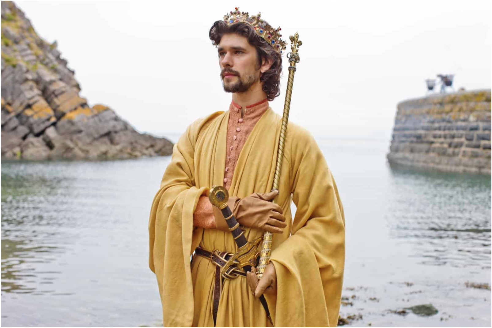 Richard stands in front of the sea, behind him, while he faces left of the camera. He holds an ornate gold sceptre that goes up to his head, and wears pink and gold robes. A sheathed sword is fastened to his brown and gold leather belt.