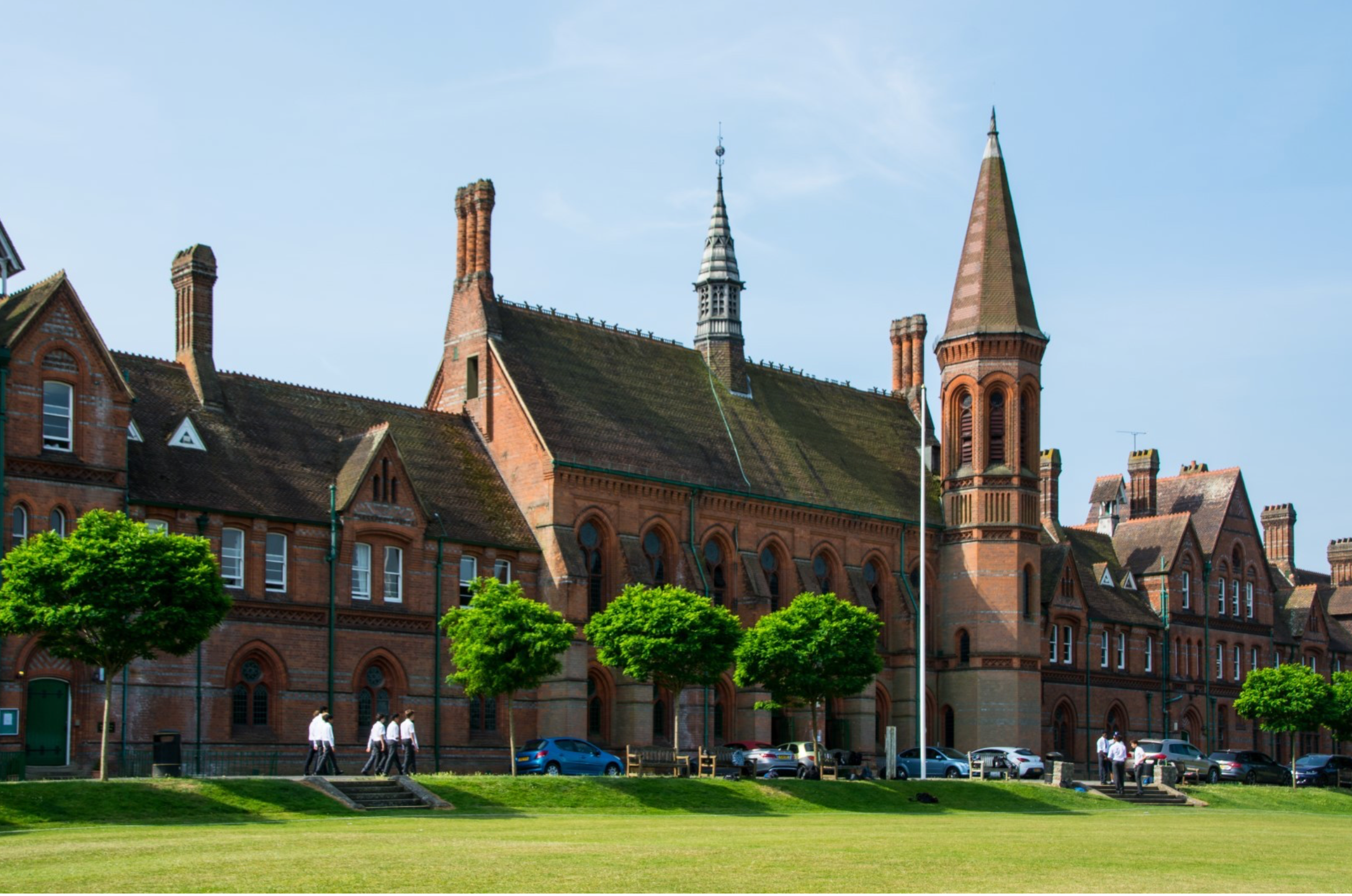 An image of Reading School. The building has dark red bricks, and is ornate with 2 spires, a vaulted chapel, and a tower. Cars are parked in front of it with white shirted students walking there too. A lawn can be seen in the foreground.