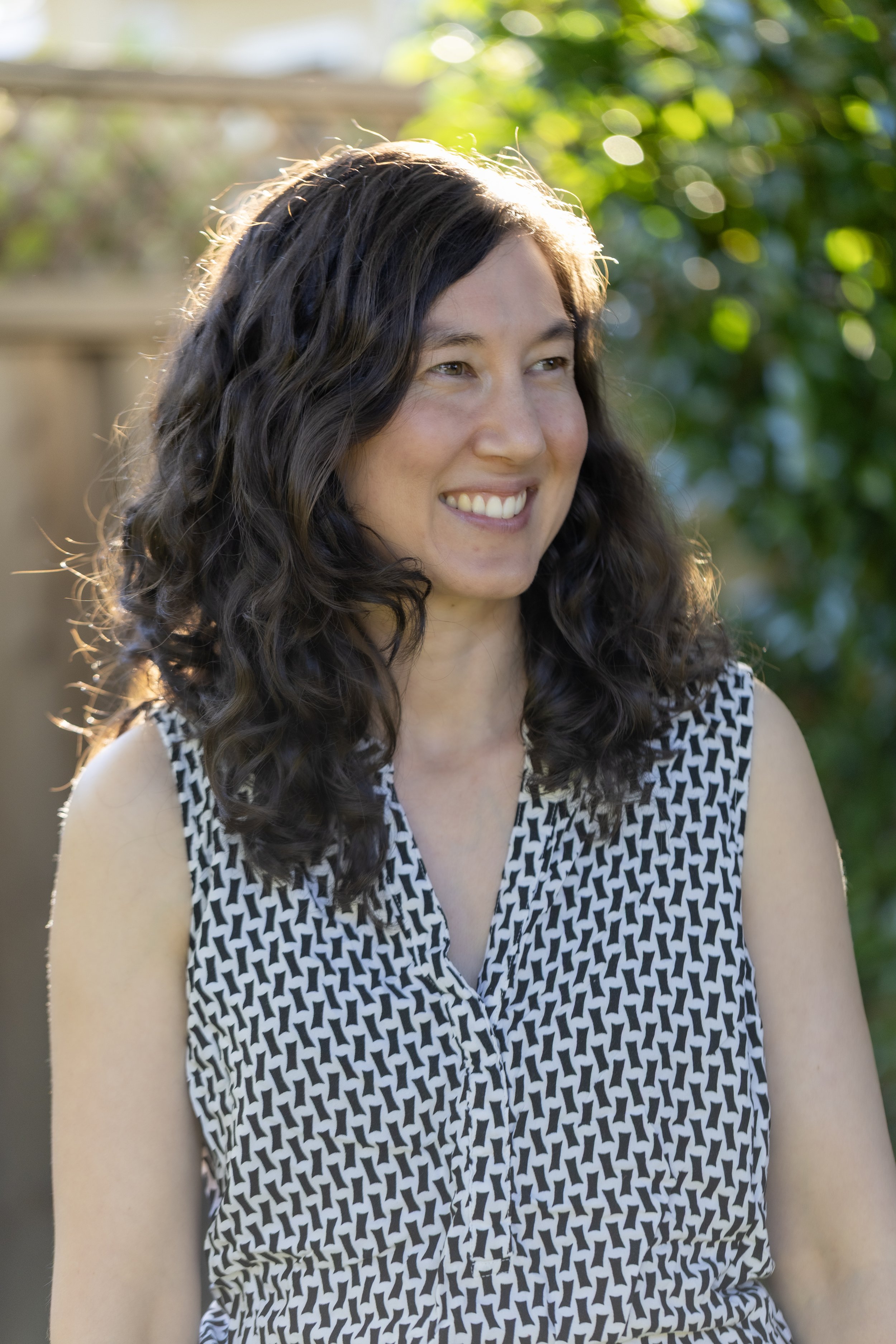 A woman with shoulder-length curly dark hair, wearing a sleeveless black and white patterned dress, smiling outdoors in sunlight with blurred green foliage in the background.