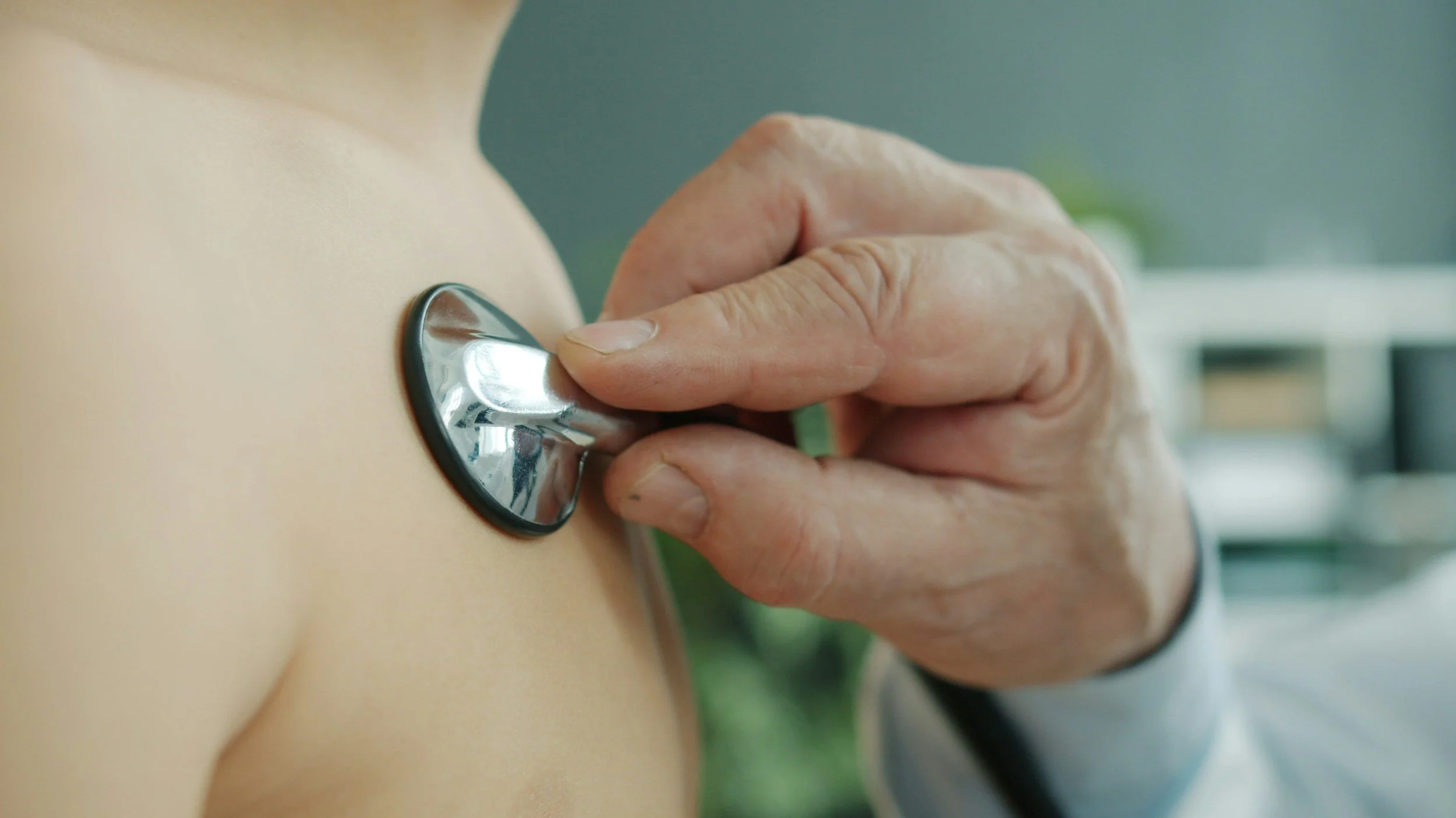 Close-up of a healthcare professional's hand inserting a stethoscope's chest piece onto a patient's bare chest.