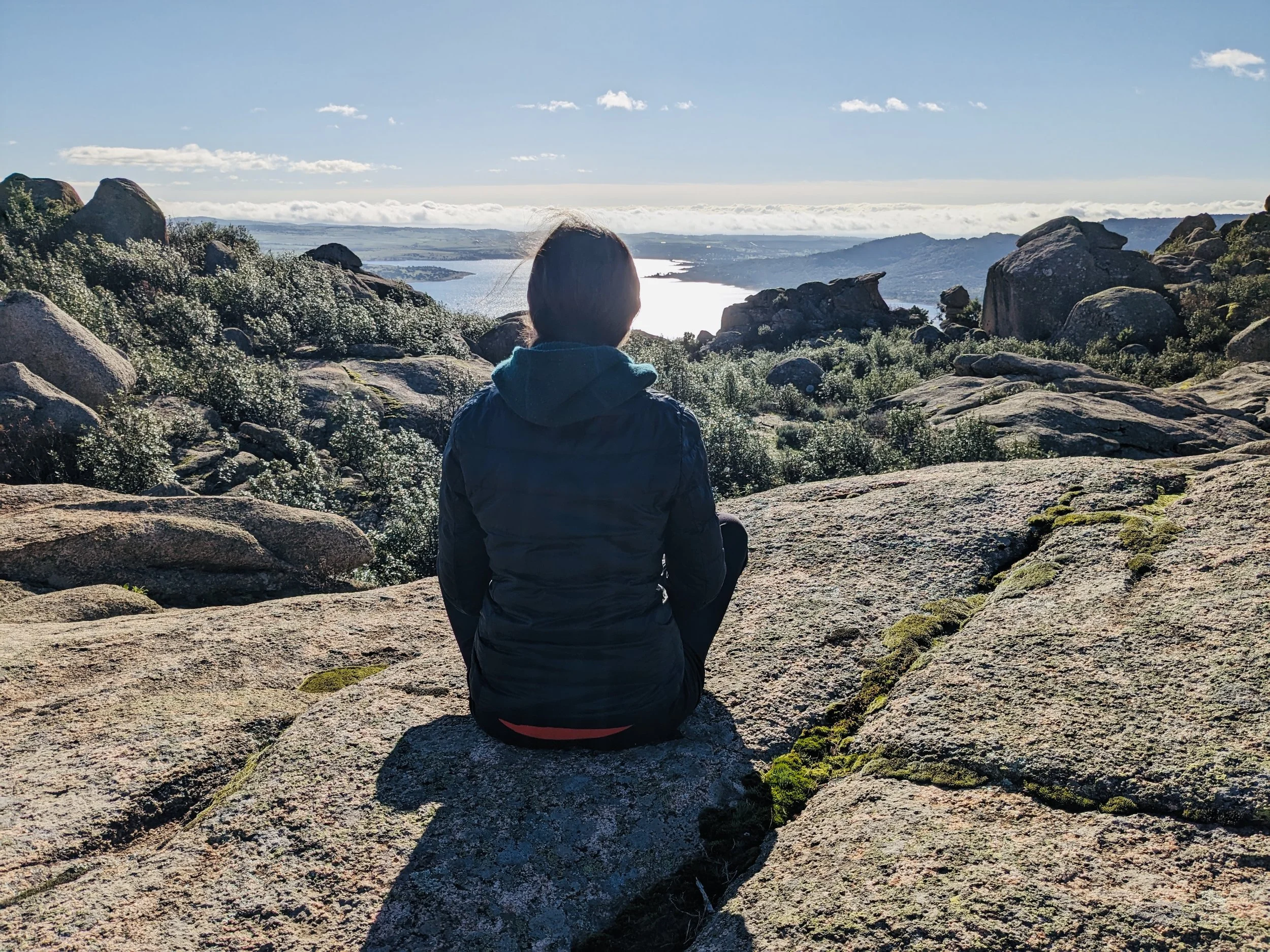 Person sitting on a rocky hilltop overlooking a lake and distant mountains on a clear day