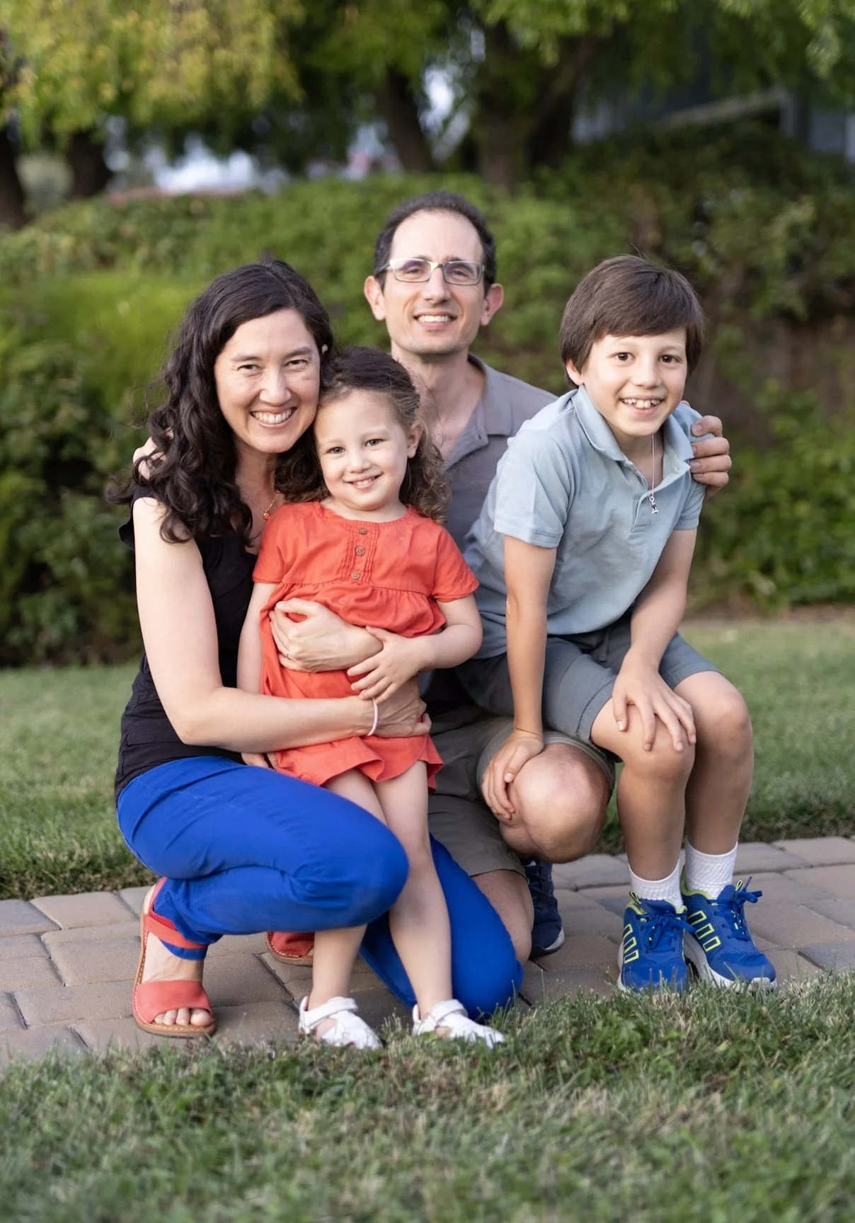 Family of four posing outdoors on a lawn with trees in the background. The mother has dark curly hair and is wearing a black top and blue pants. The father has short dark hair, glasses, and is wearing a gray shirt. The daughter has curly hair, is wearing a red dress, and is sitting in her mother's lap. The son has straight dark hair, is wearing a light blue polo and shorts, and is crouching with one knee on the ground.