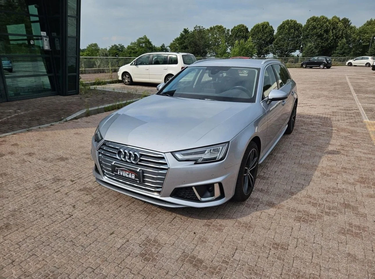 Carro prata da Audi estacionado em um pátio, com outros veículos ao fundo e árvores ao redor.