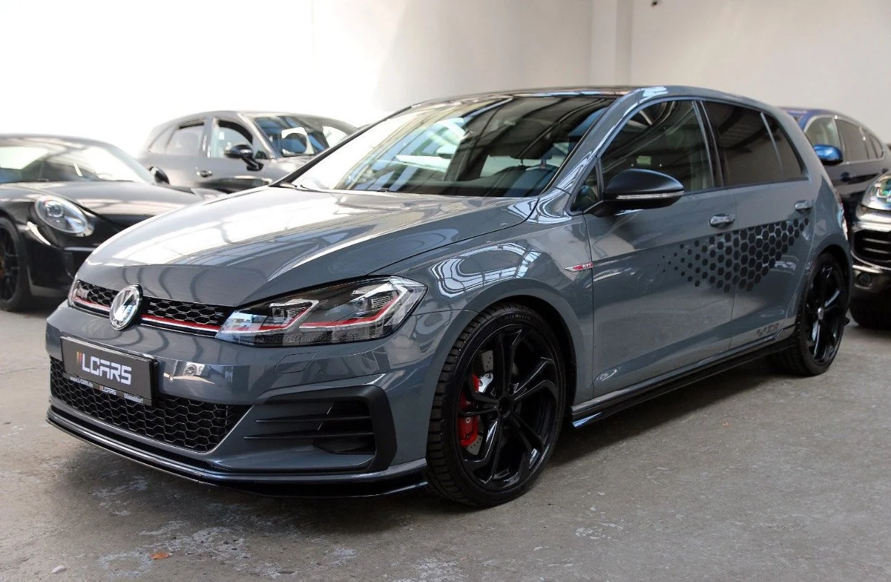 Carro hatchback cinza com detalhes pretos, rodas esportivas pretas e freios vermelhos, estacionado em uma garagem com outros carros ao fundo.