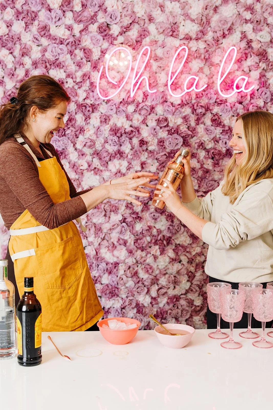 Two women laughing while preparing drinks at a party, with a pink flower wall and neon sign that says 'Oh La La' behind them.
