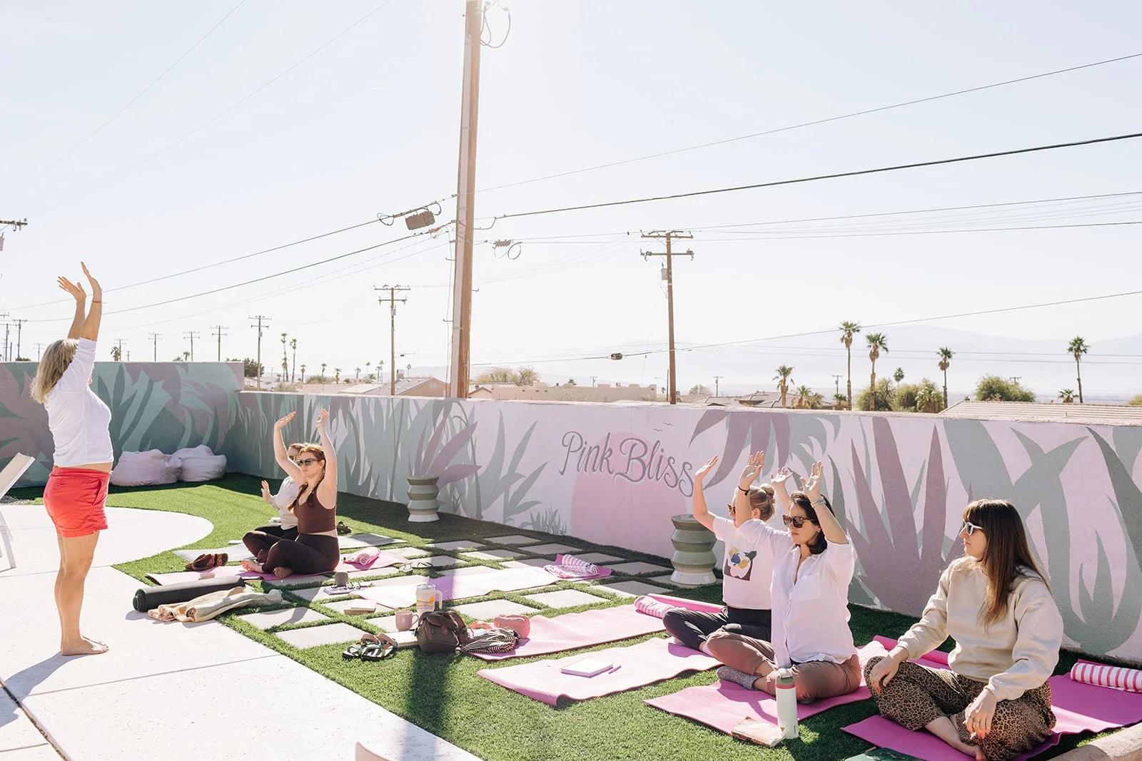 Group of women participating in a yoga or meditation class outdoors on pink mats. An instructor is standing and leading the session, while the women sit cross-legged and raise their arms. The setting is a rooftop with a painted wall that reads 'Pink