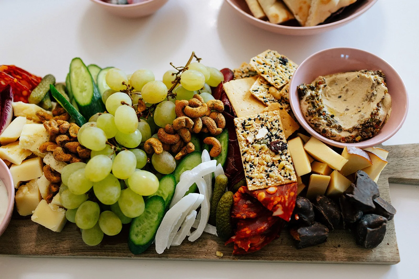A cheese and fruit board with grapes, melon, cucumber, nuts, cheeses, crackers, dried meats, and various spreads in bowls.
