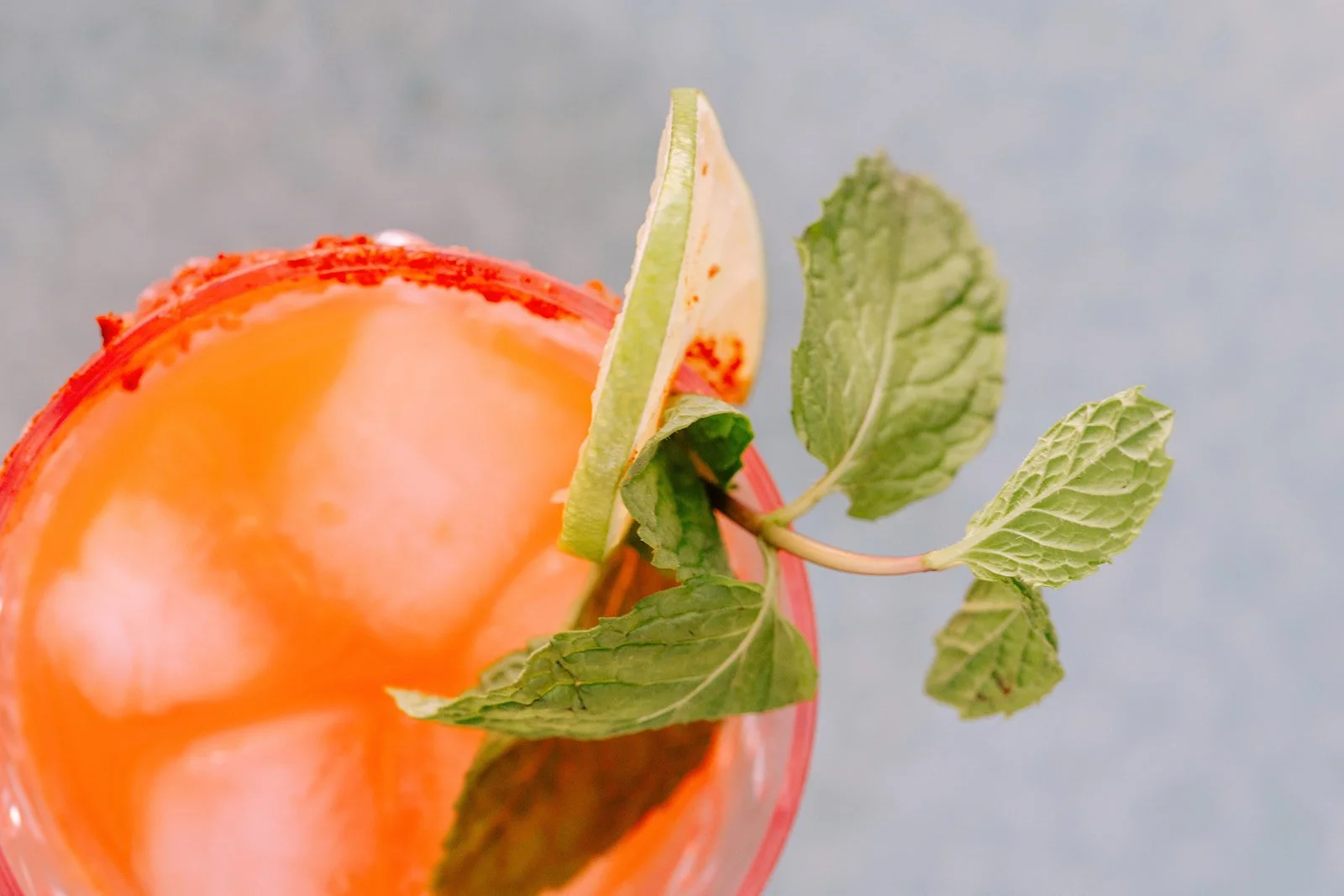 Close-up view of an orange-colored drink with a slice of lime and fresh mint leaves garnishing the rim against a light blue background.