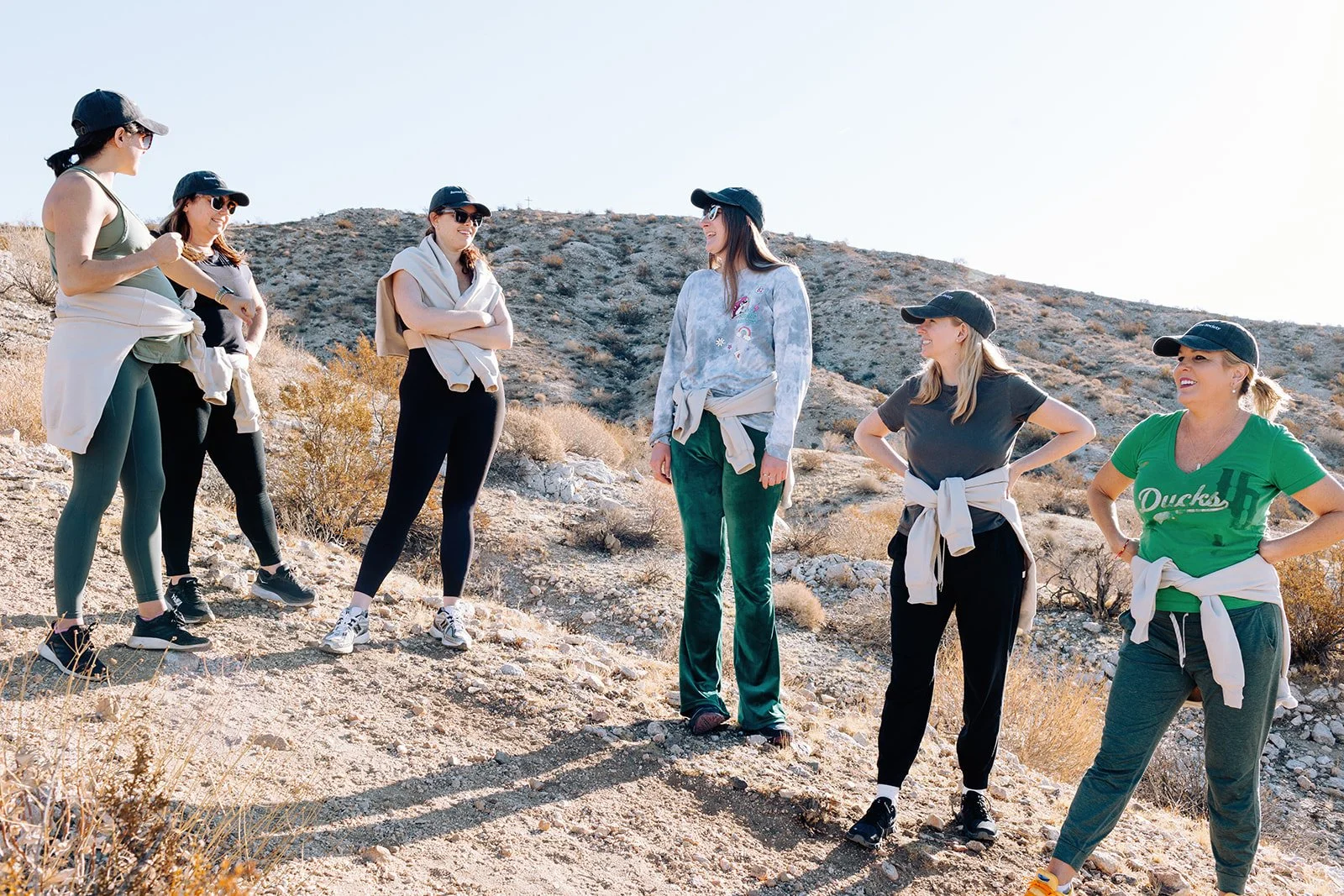 Six women standing outdoors on a trail in a desert landscape, wearing casual athletic clothing and black caps, engaging in conversation.