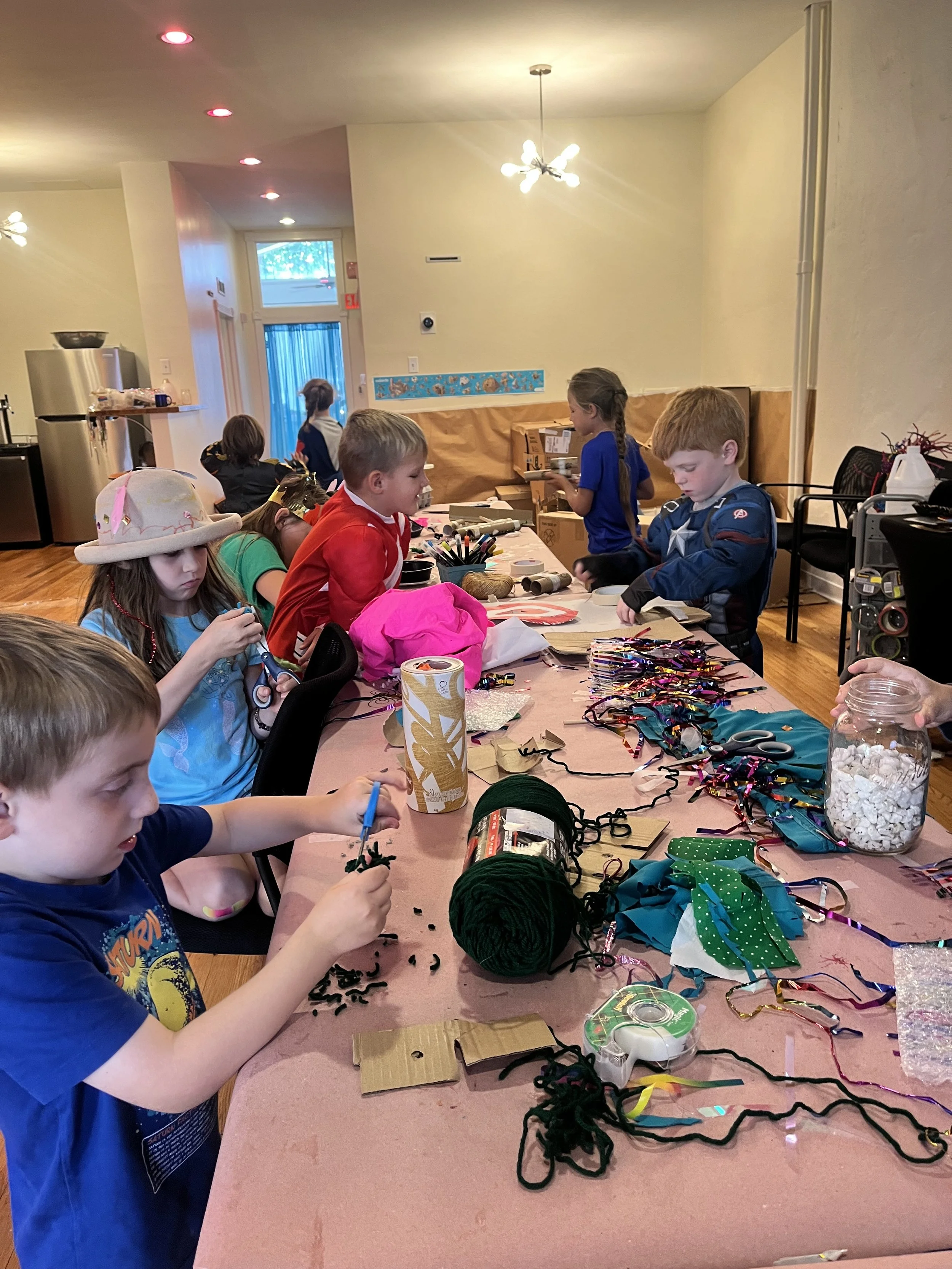 Children engaged in arts and crafts at a long table, with supplies including yarn, ribbons, scissors, and decorations, inside a well-lit room with wooden floors and minimal wall decoration.