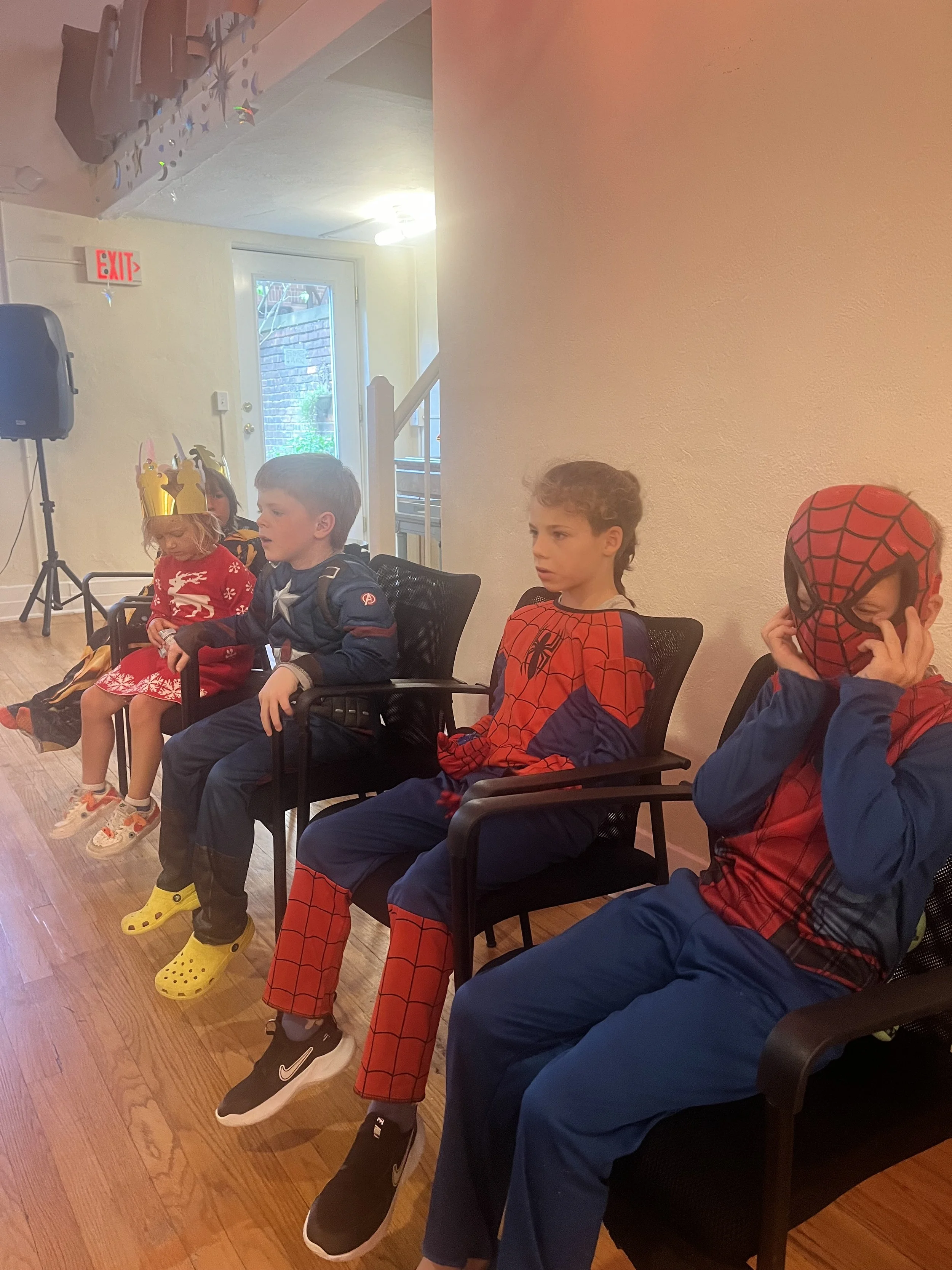 Children dressed in costumes, including Spider-Man, Dorothy, and a princess, sitting on chairs during a party or event.