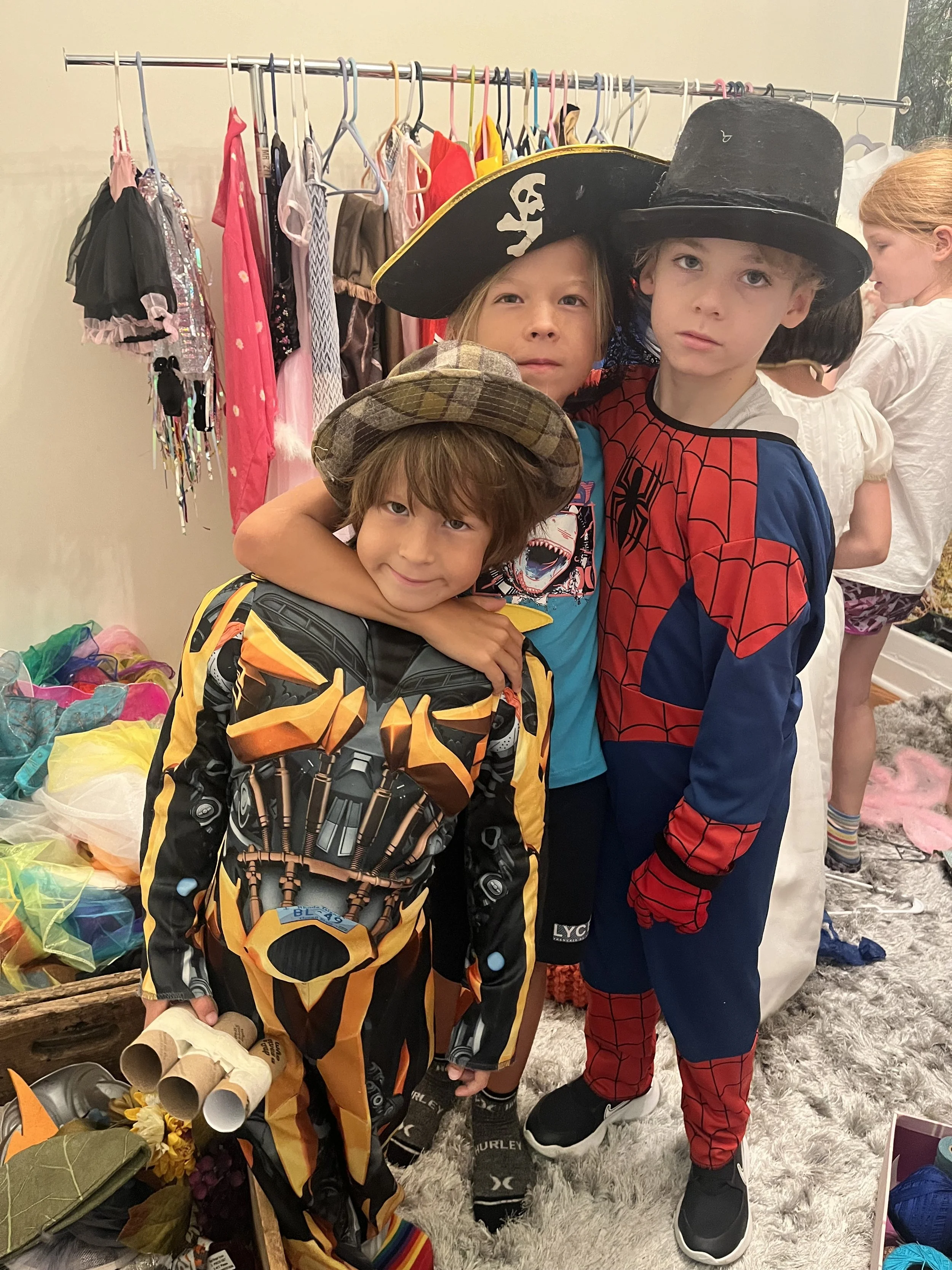 Three children dressed in costumes, including a pirate, Spider-Man, and a character from Transformers, standing together at a costume party or Halloween event.