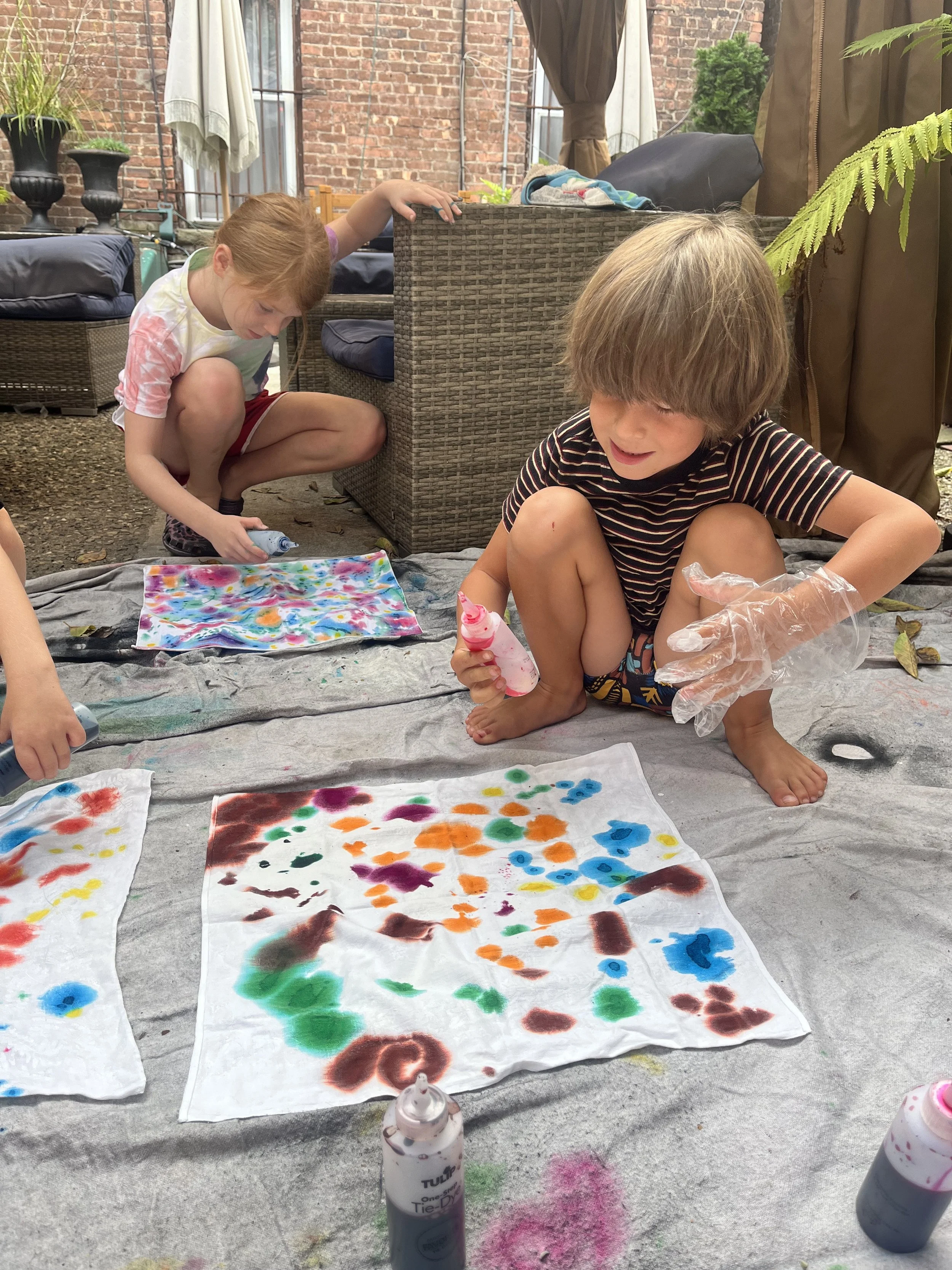 Children engaging in tie-dye activity outdoors, with colorful fabric and spray bottles on a tarp.