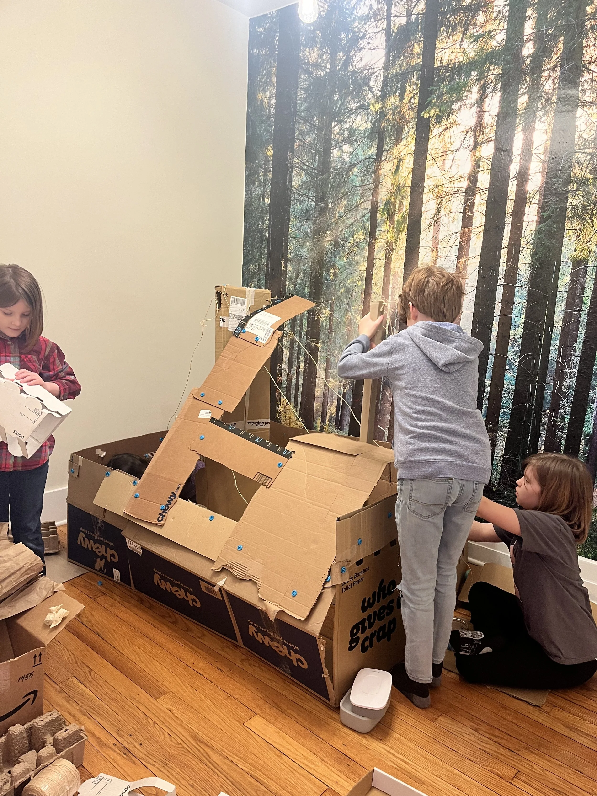 Three children building a cardboard structure inside a room with a forest mural on the wall.