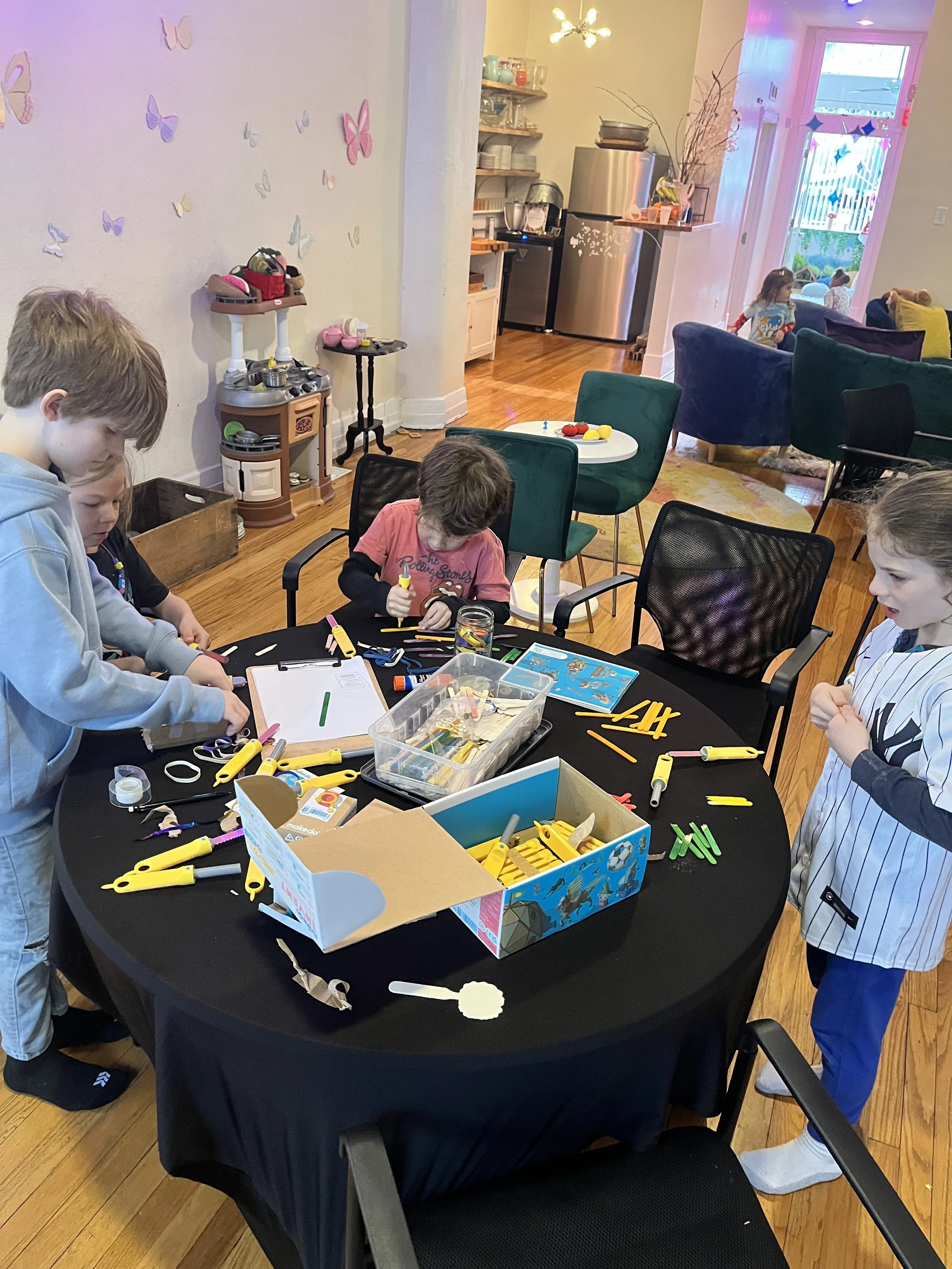 Children gathered around a black table engaged in arts and crafts, with craft supplies scattered on the table. The room has colorful butterfly wall decals, a small play kitchen, and a cozy living area in the background.