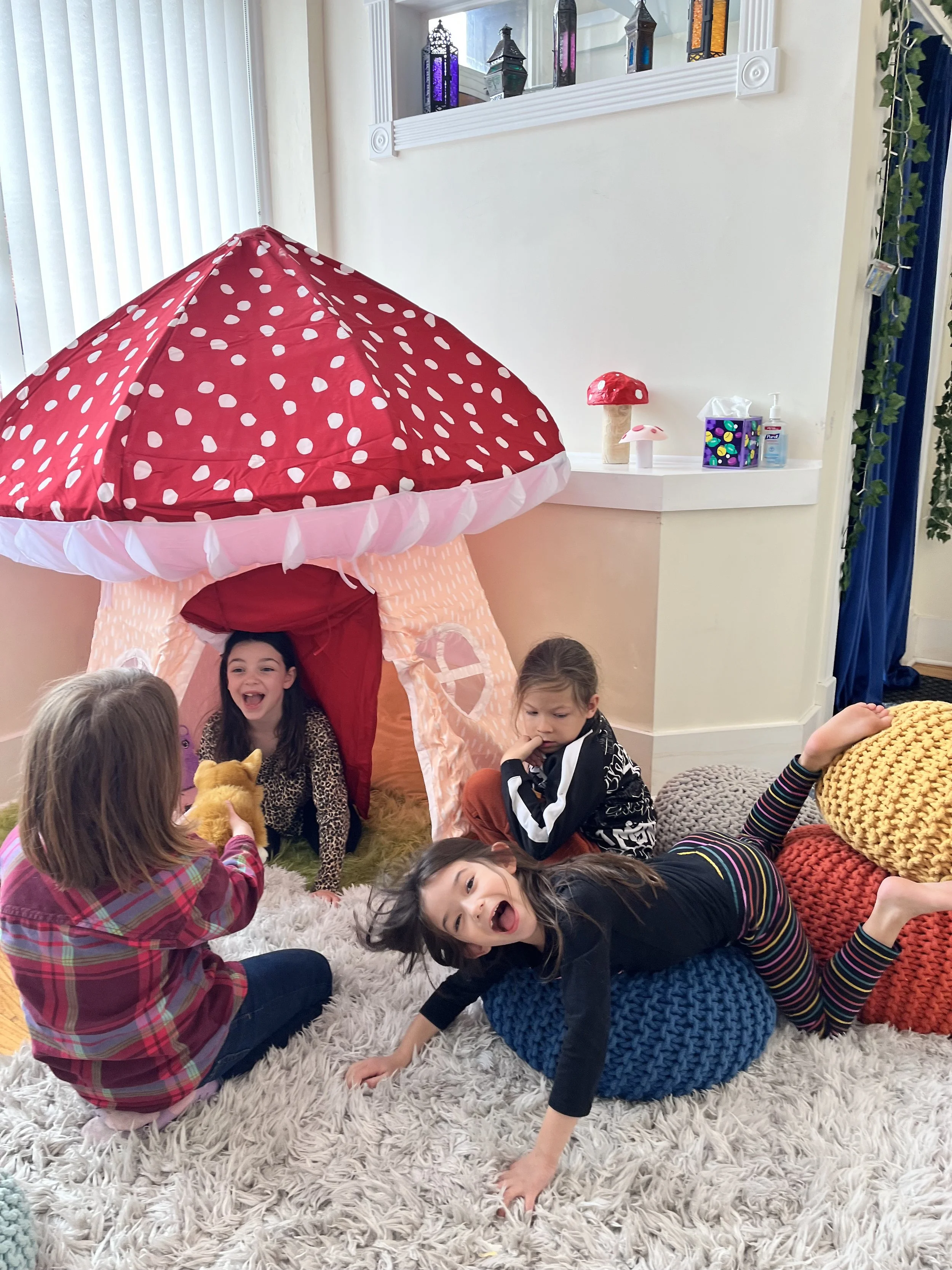 Four children playing around a red and white mushroom-shaped play tent in a cozy room with colorful pillows, plush carpet, and decorative items on a shelf.