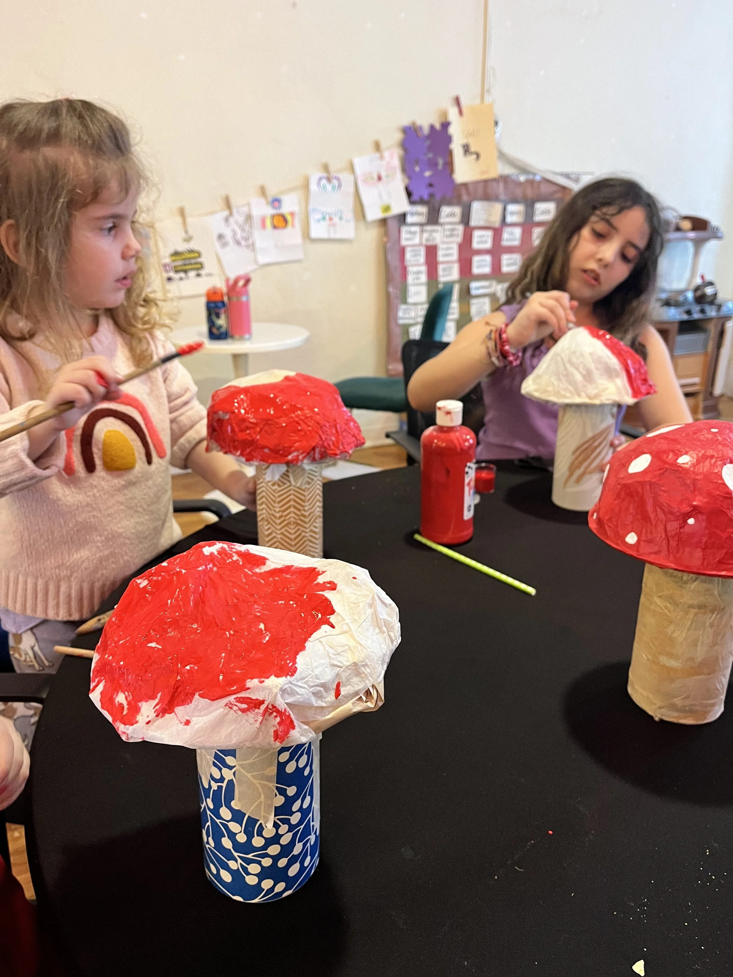 Two young girls painting paper mushroom craft decorations with red paint, sitting at a black table in a classroom.
