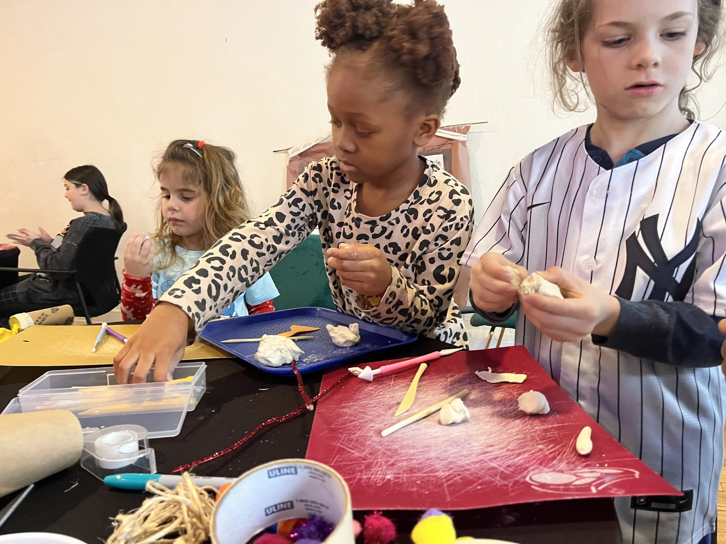 Children engaging in a craft activity, working with white modeling clay and craft tools at a table with various supplies around.