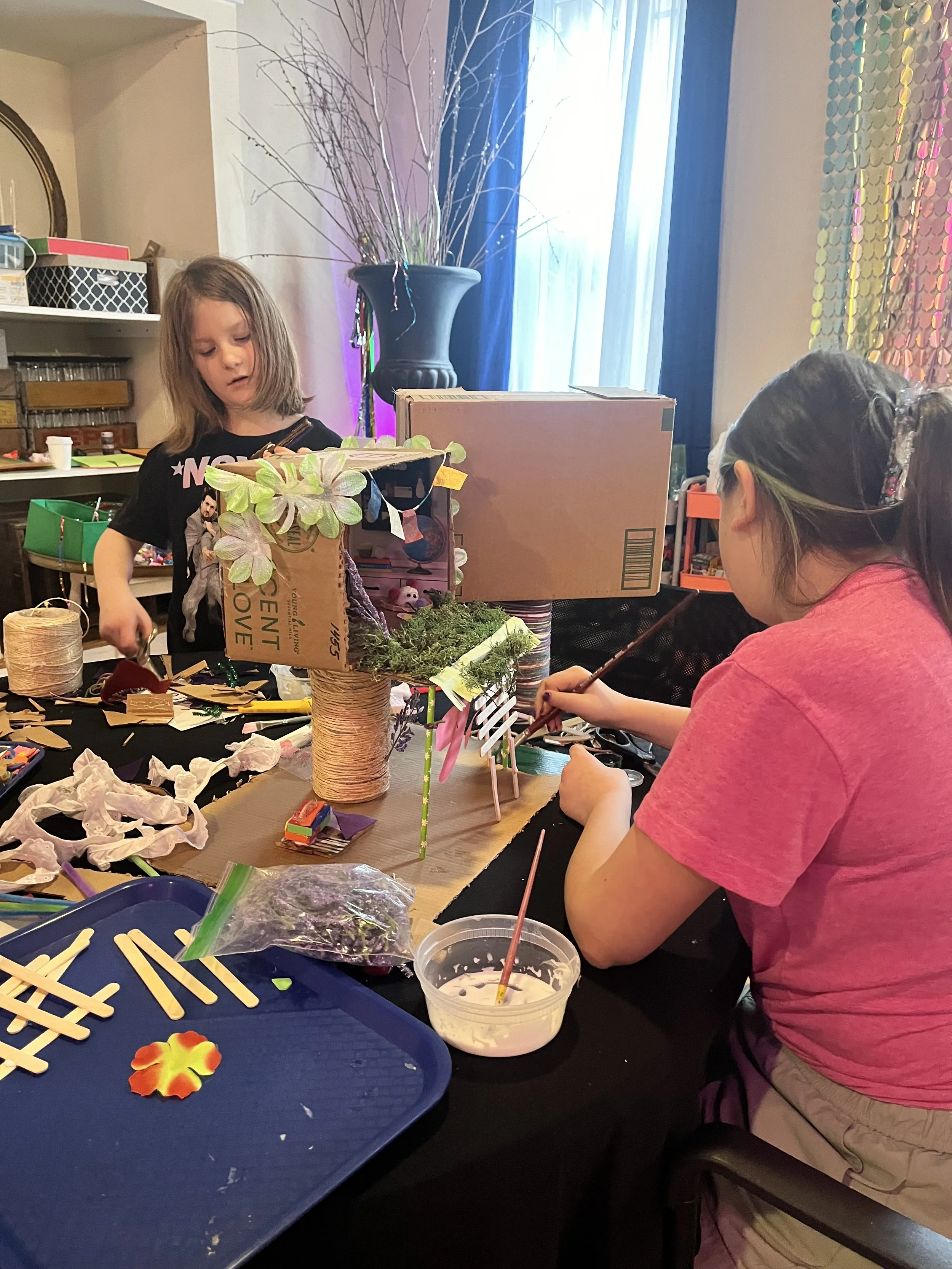 Two children craft a decorative plant with a cardboard box, twine, and artificial foliage at a table cluttered with craft supplies.