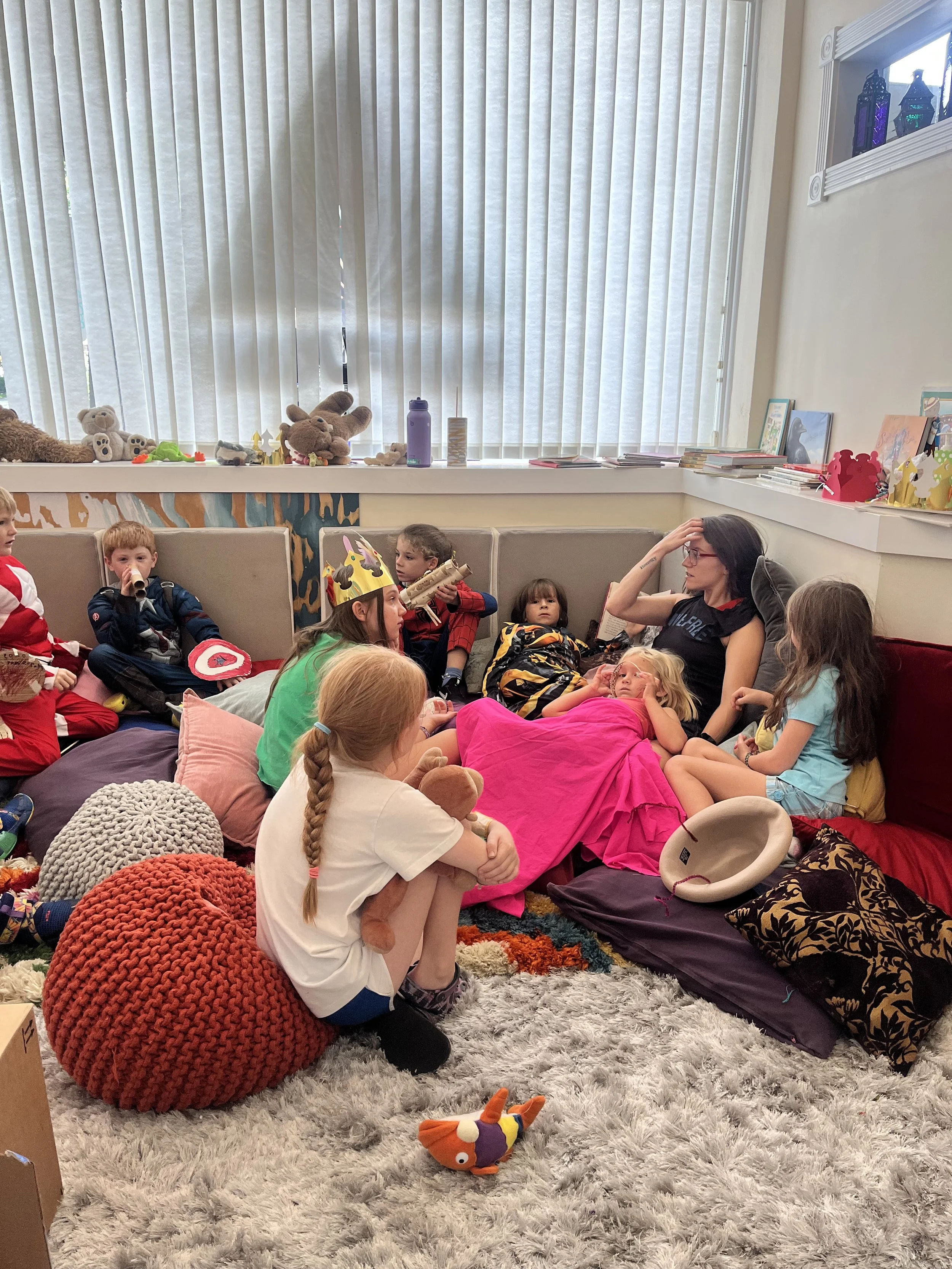 Children and an adult resting on colorful cushions in a cozy room with stuffed animals and toys on a ledge.