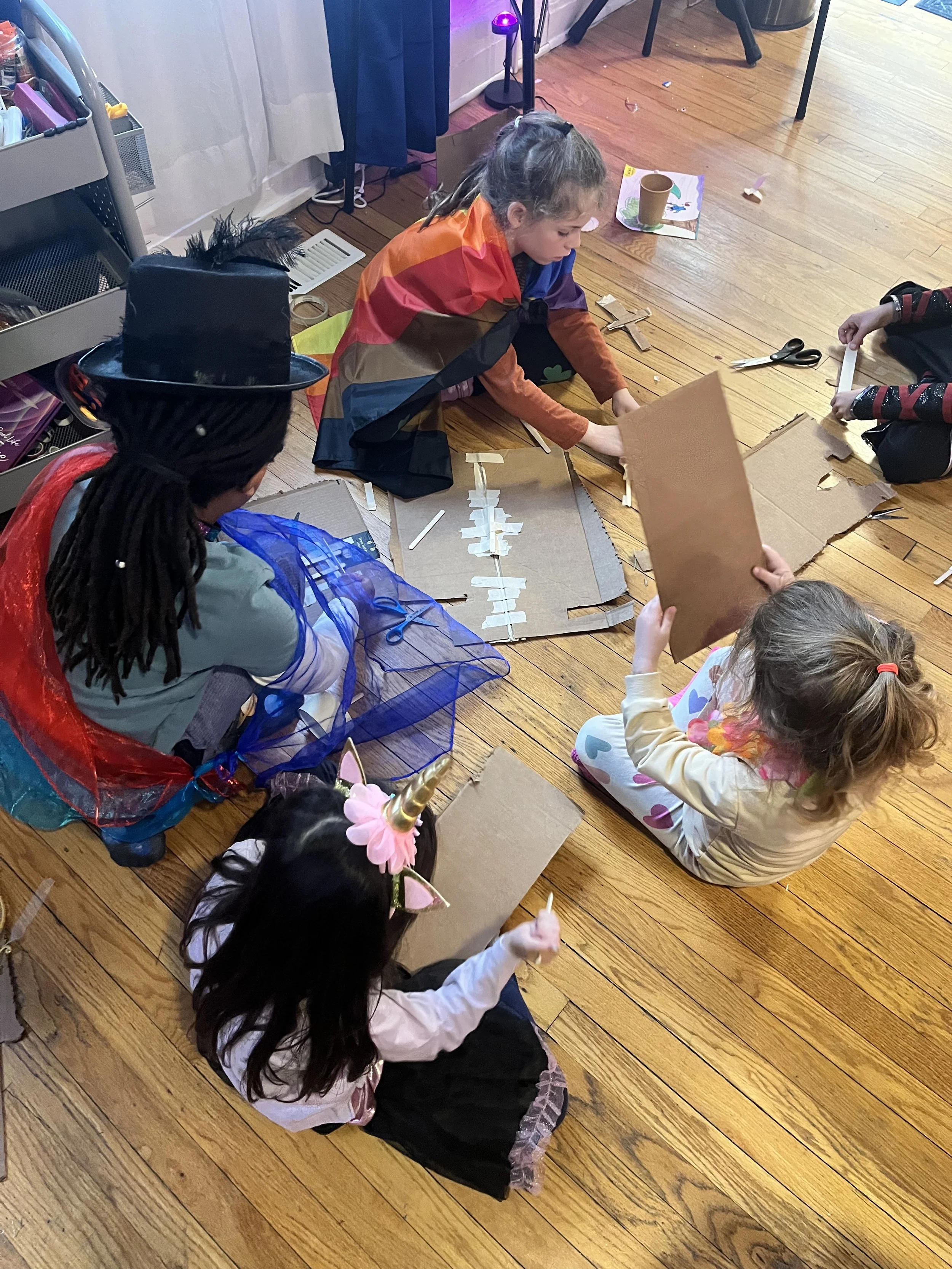 Children dressed in costumes, including a unicorn headband and witch outfit, are sitting on the wooden floor working on cardboard craft projects.