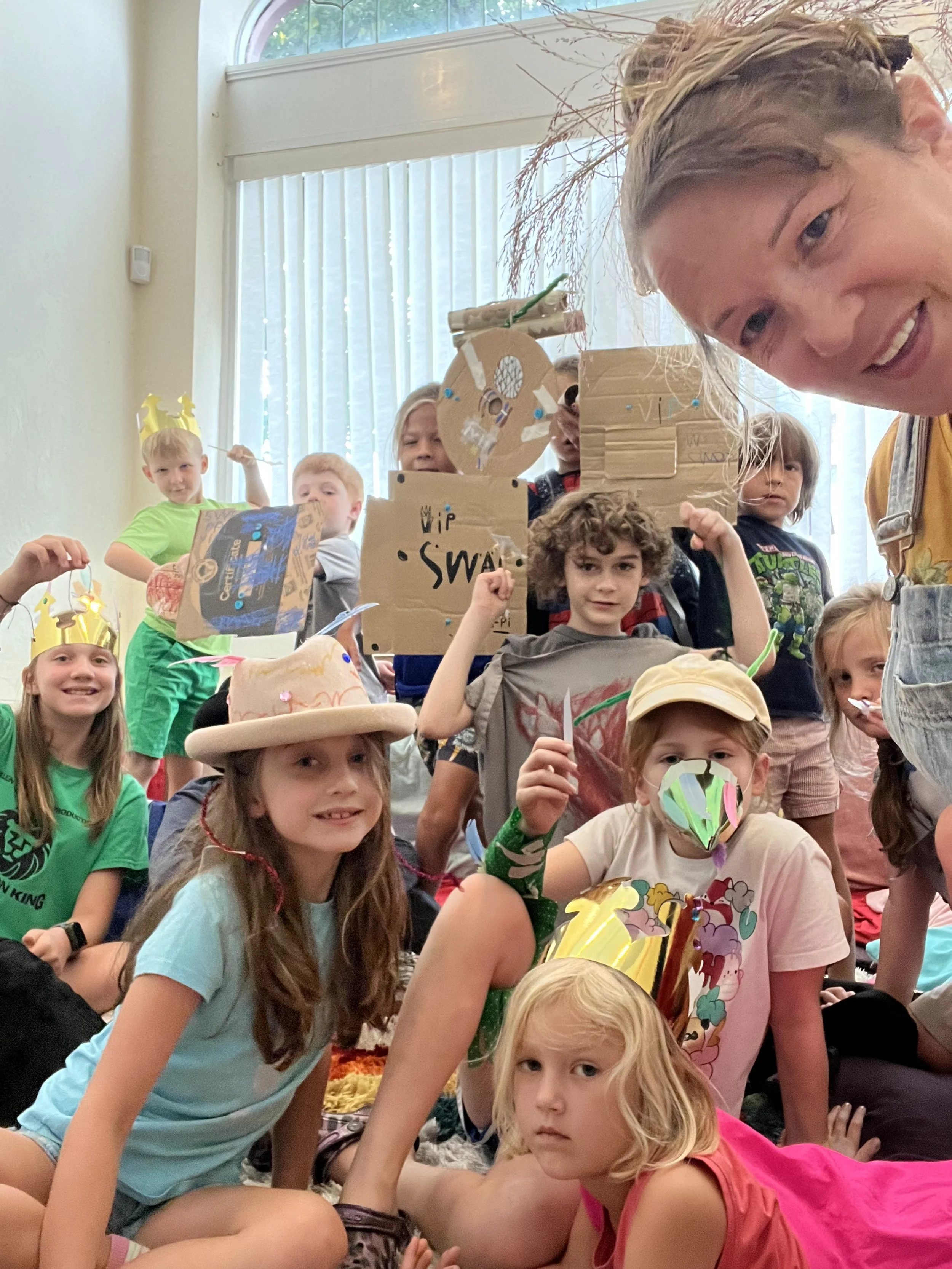 A group of children at a birthday party, wearing paper crowns, holding signs, and wearing birthday hats and masks, with a woman taking a selfie with them in a brightly lit room.