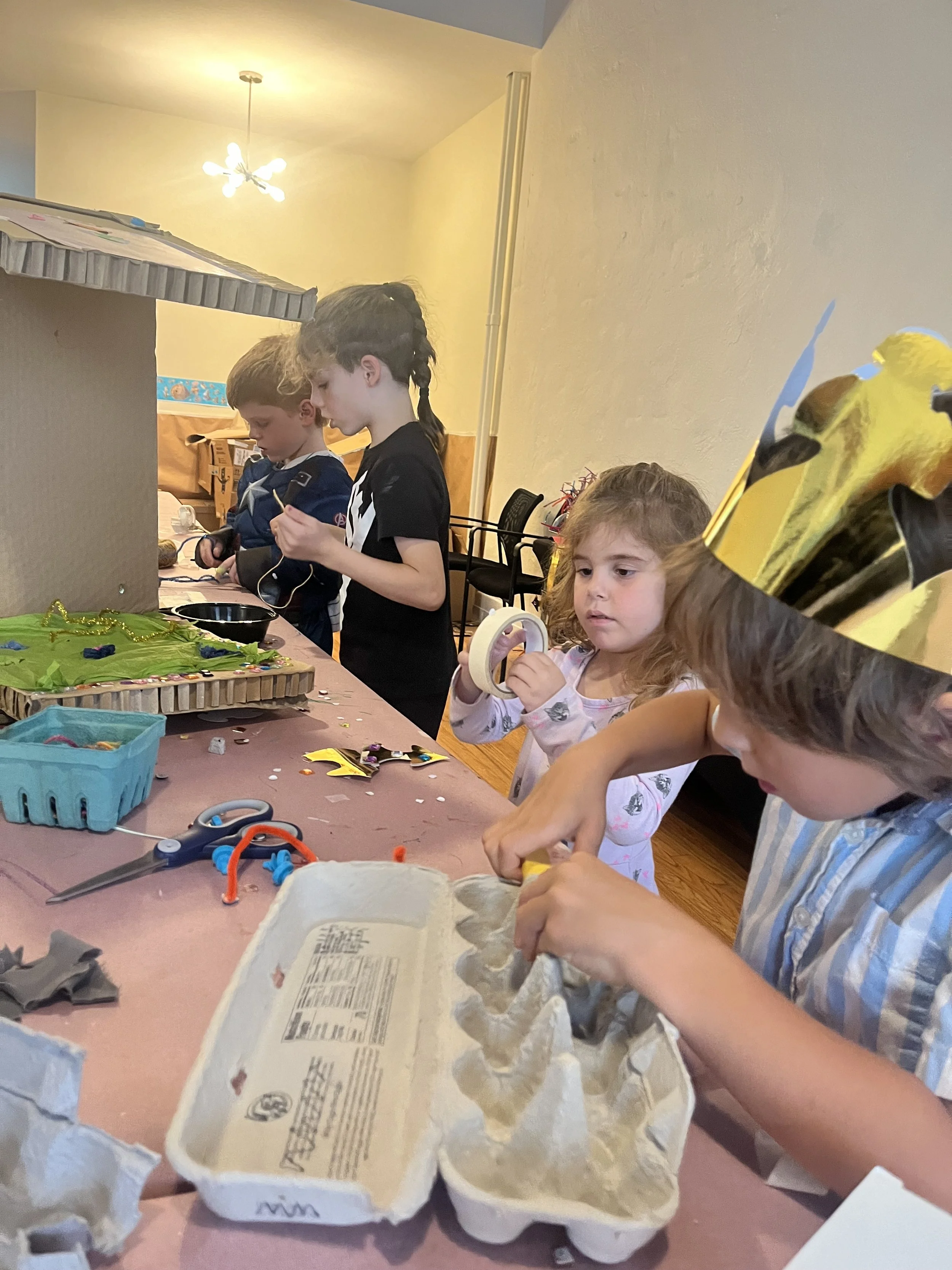 Children decorating and working on arts and crafts at a table, with various craft supplies like tape, scissors, and egg cartons in a room with a beige wall and overhead lighting.