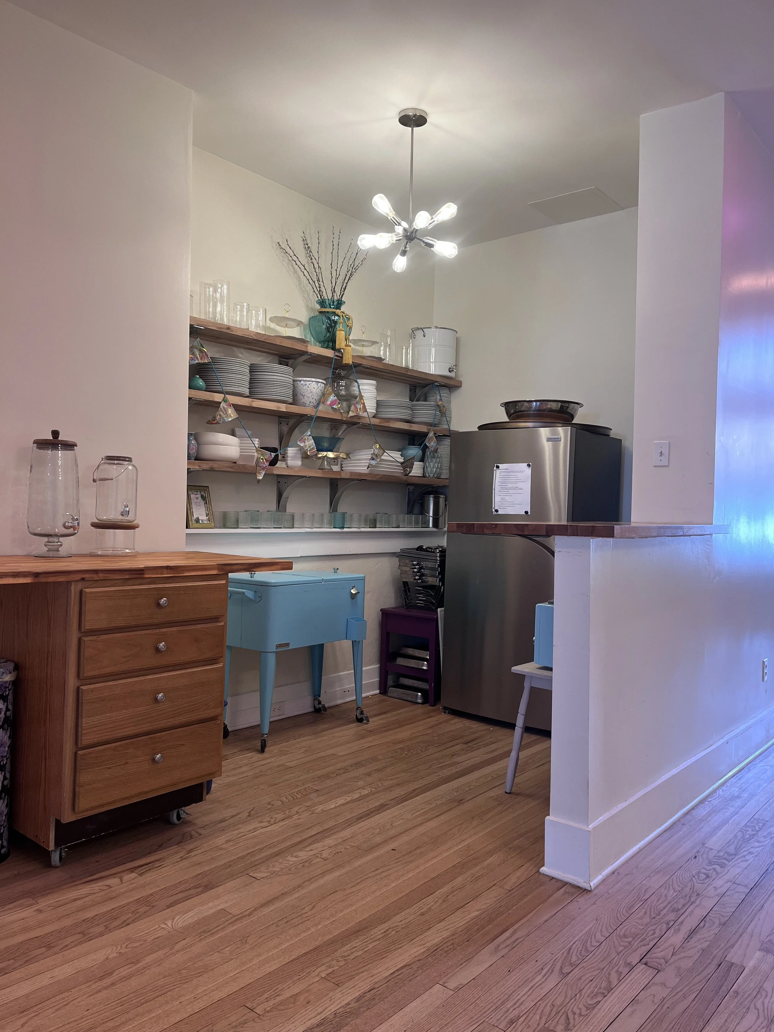 Open kitchen shelf with plates, bowls, glassware, and colorful cups, coffee machine, and utensil jar.