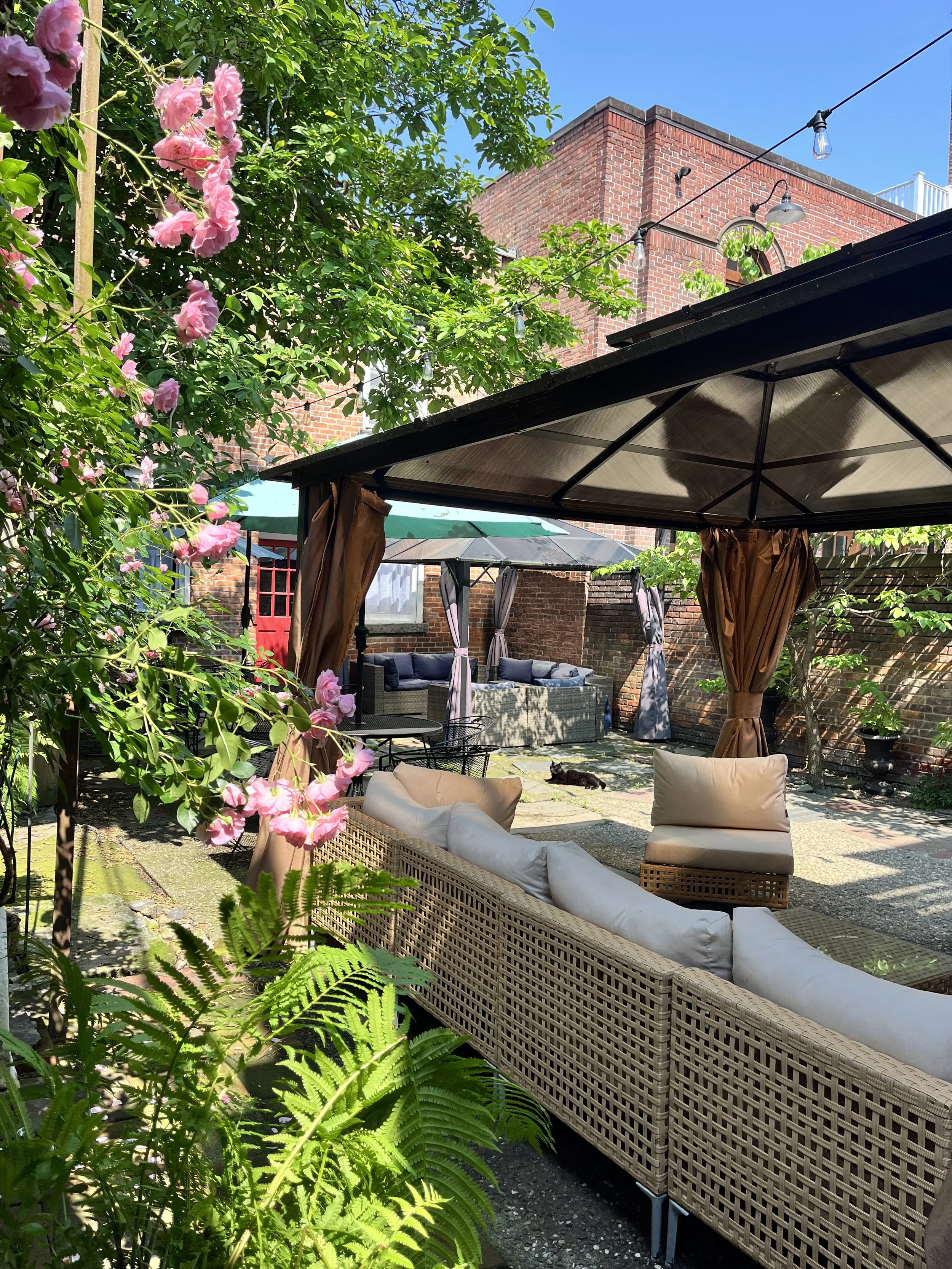 Outdoor patio with black metal tables and chairs, decorated with woven placemats, green glasses, and cloth napkins tied around the placemats, under blue and white patio umbrellas, with string lights and lush green plants in the background.