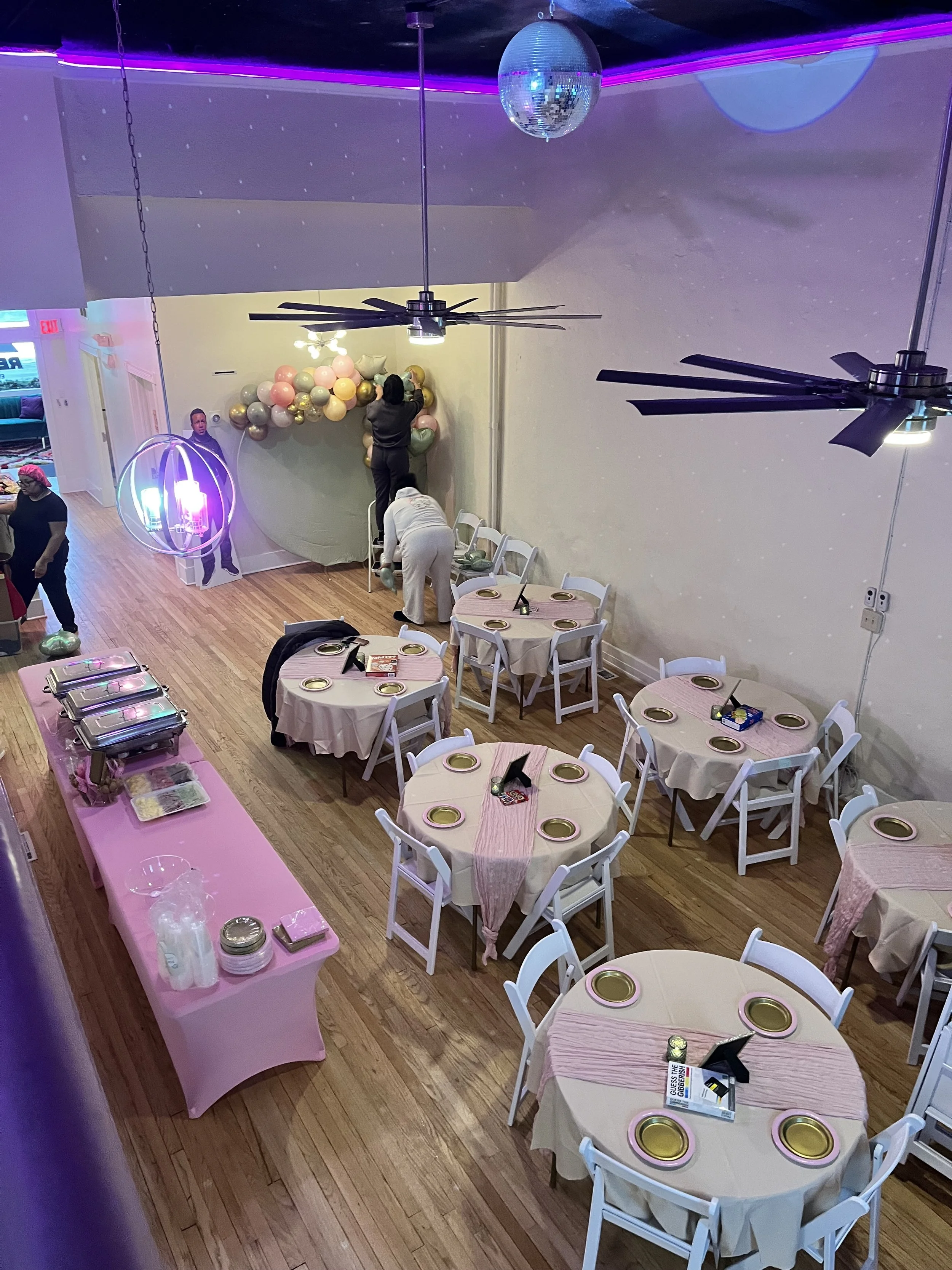 Interior of a decorated event space with round tables covered in blue and black tablecloths, black chairs, a black lounge with yellow pillows and green arch decor, ceiling fans, and a wall with string lights and a door to outside.