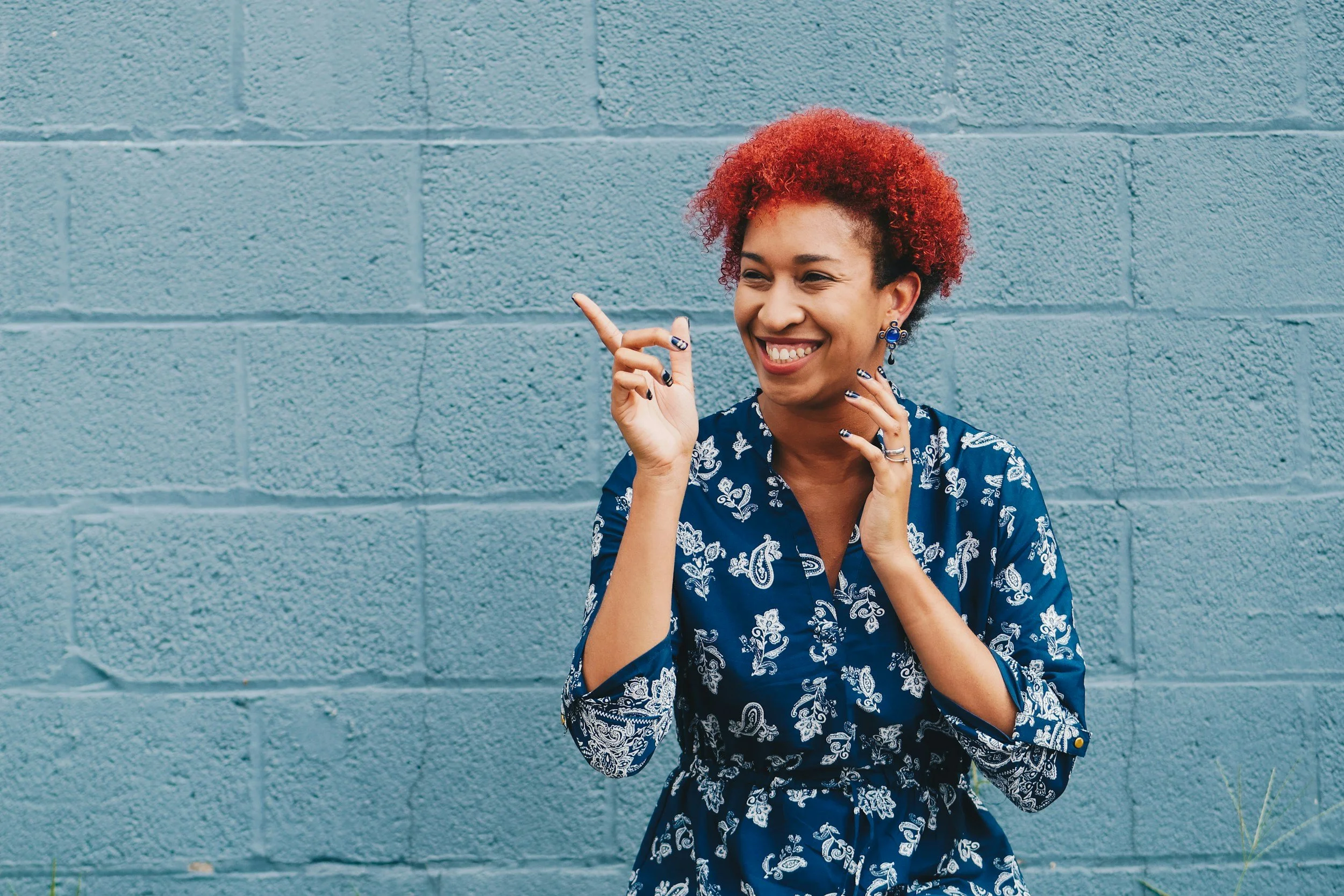 A woman with short curly red hair smiling and pointing to her left while standing in front of a blue brick wall.