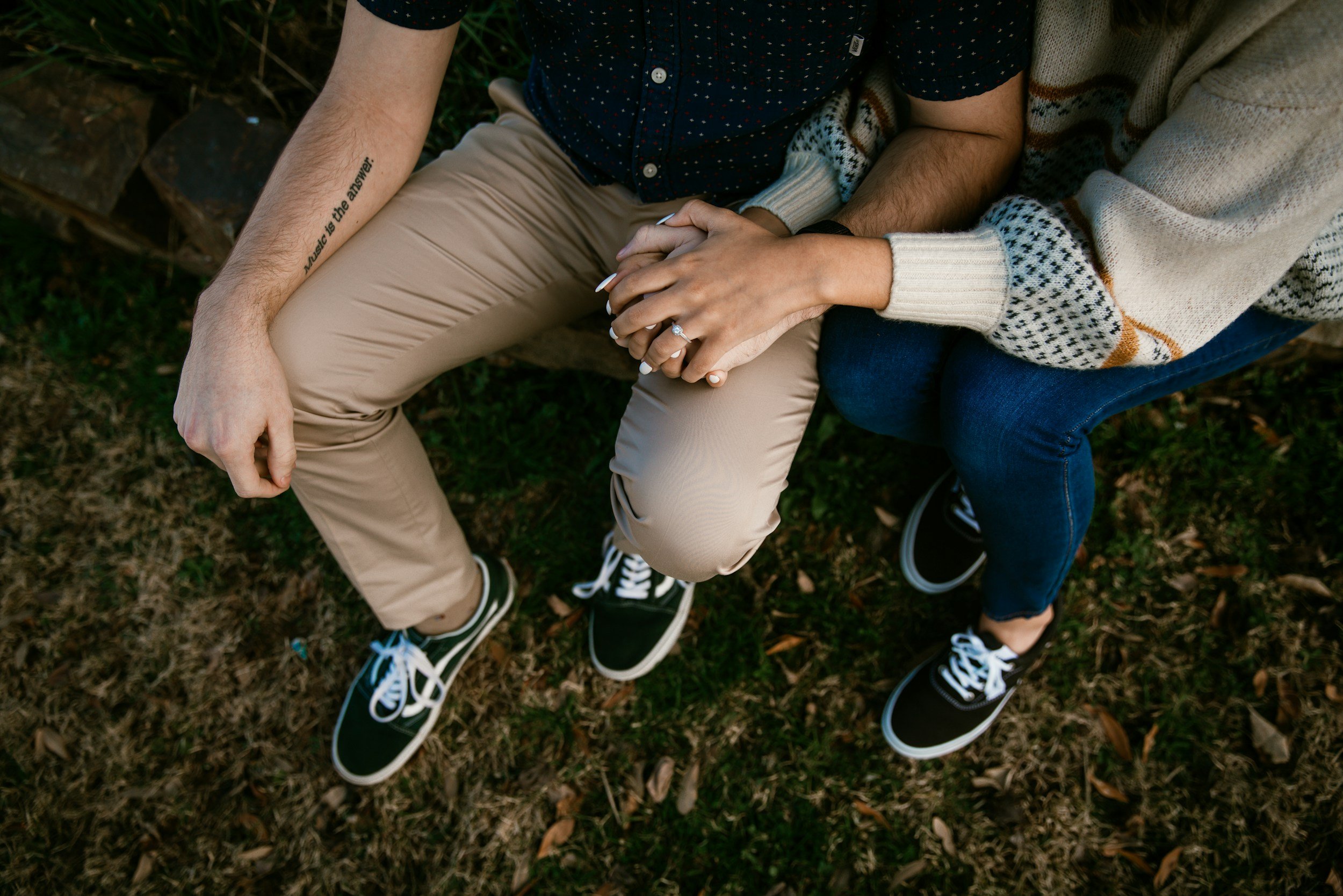 A couple sitting outdoors on a wooden bench, holding hands, viewed from above. The man is wearing beige pants, a dark shirt with small white dots, and black sneakers. The woman is wearing a beige sweater with a pattern, blue jeans, and black sneakers. The woman is also wearing a ring on her finger, and there is a tattoo on the man's arm.