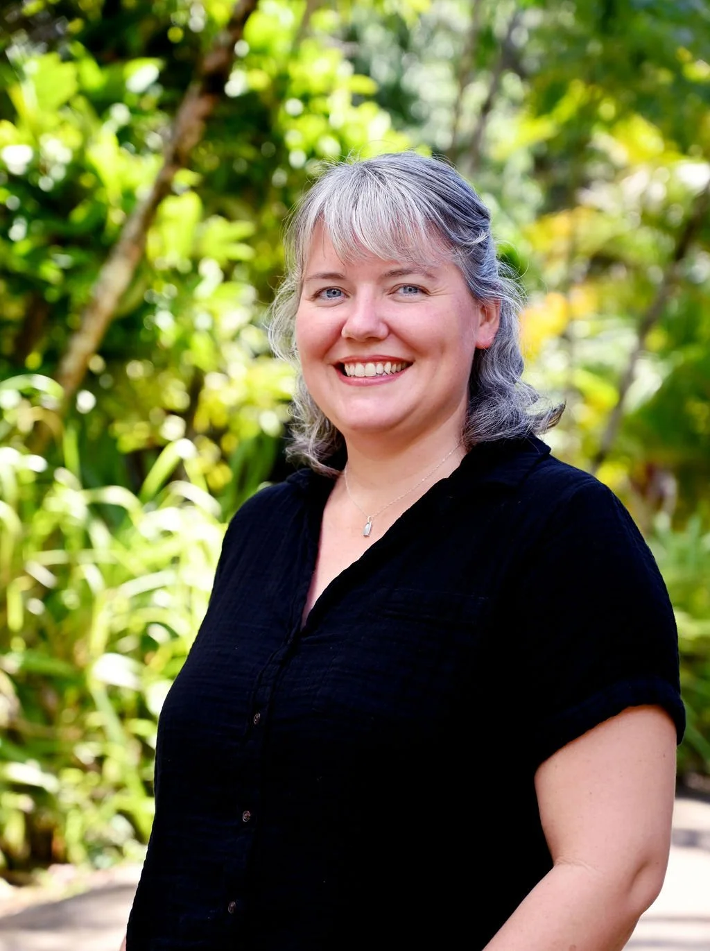 A smiling woman with gray hair and blue eyes, wearing a black top, outdoors with green foliage in the background.