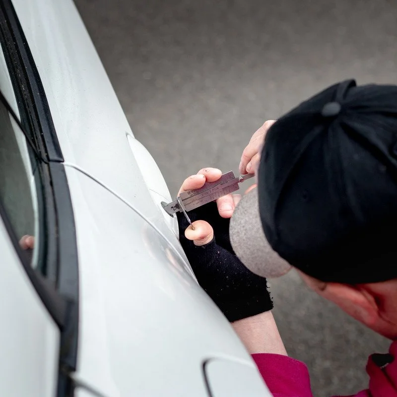 A person wearing a black cap and gray clothing uses a caliper to measure the thickness of a scratch or dent on the side of a white vehicle.
