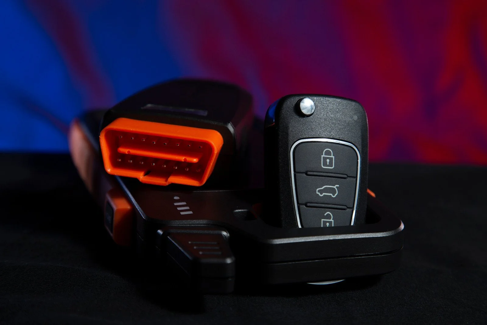Close-up of a car key fob with lock, unlock, and trunk buttons, resting in a black charging or docking station with a colorful blurred background.