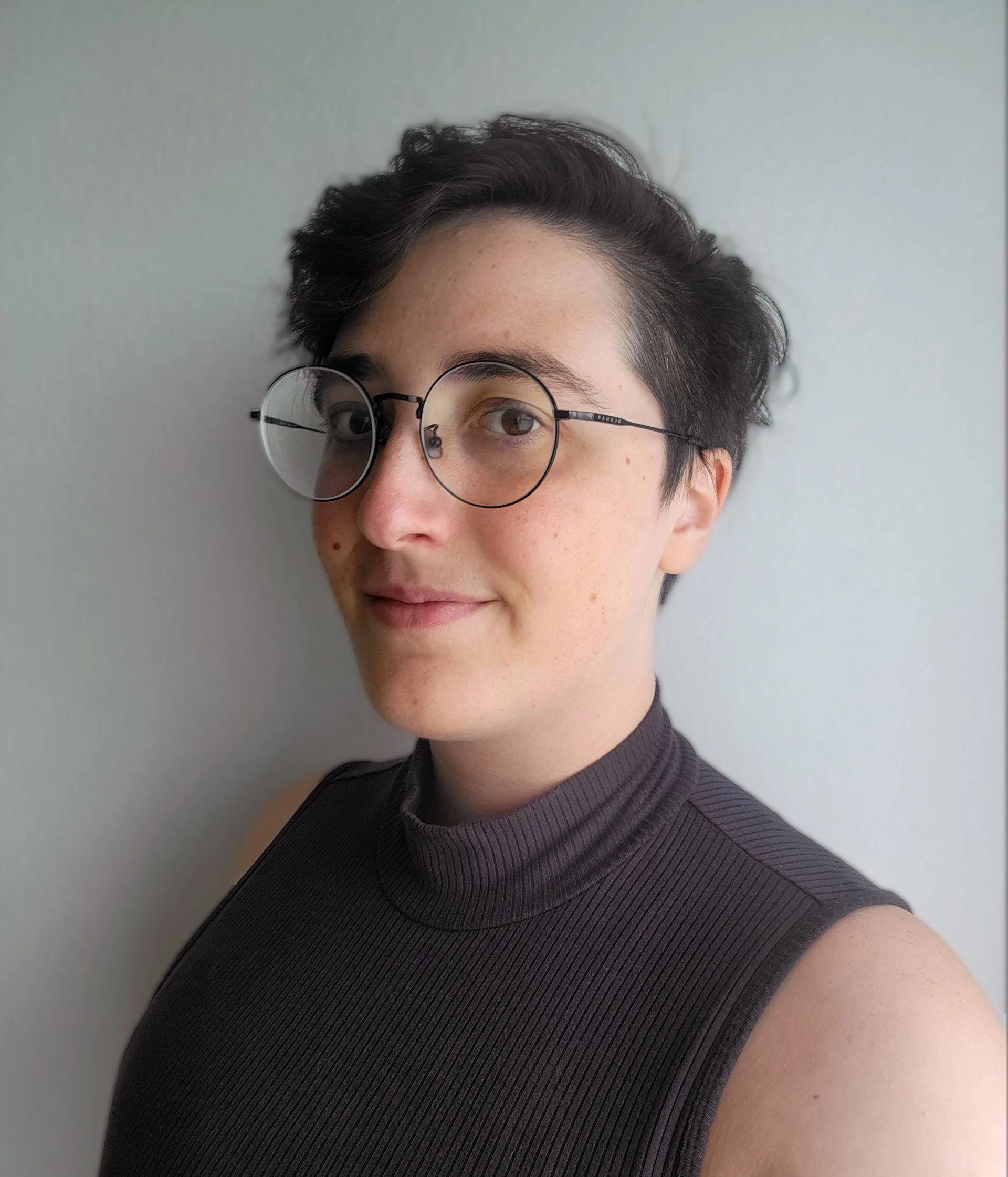 Portrait of a person with short dark hair, wearing round glasses and a sleeveless black turtleneck, standing against a plain light wall.