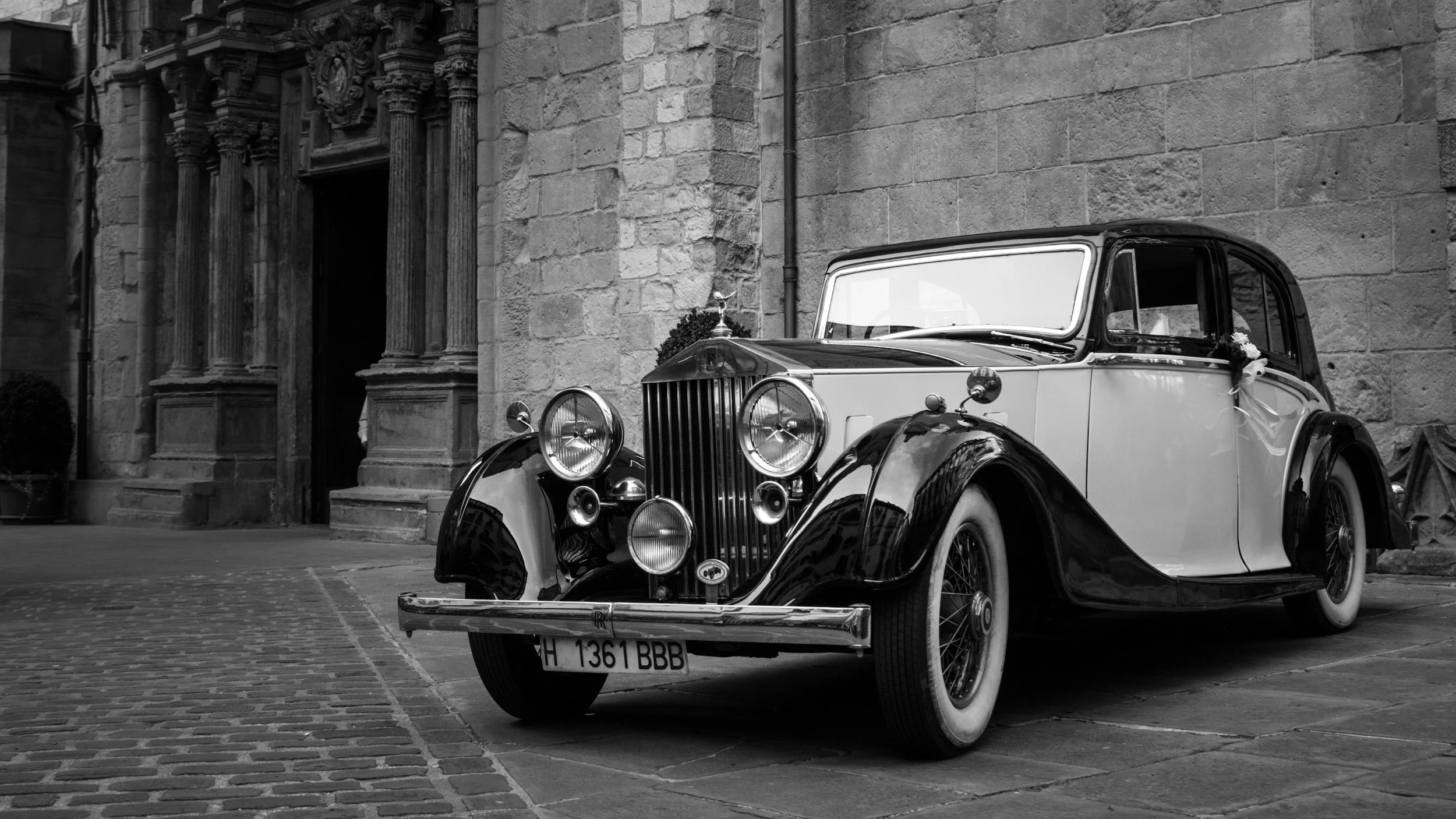 A vintage black and white luxury car parked on a cobblestone street outside a historic stone building with decorative columns and an arched doorway, with wedding decorations on the car door.