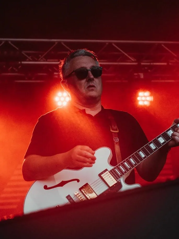 Musician wearing sunglasses playing a white electric guitar on stage with red lighting.