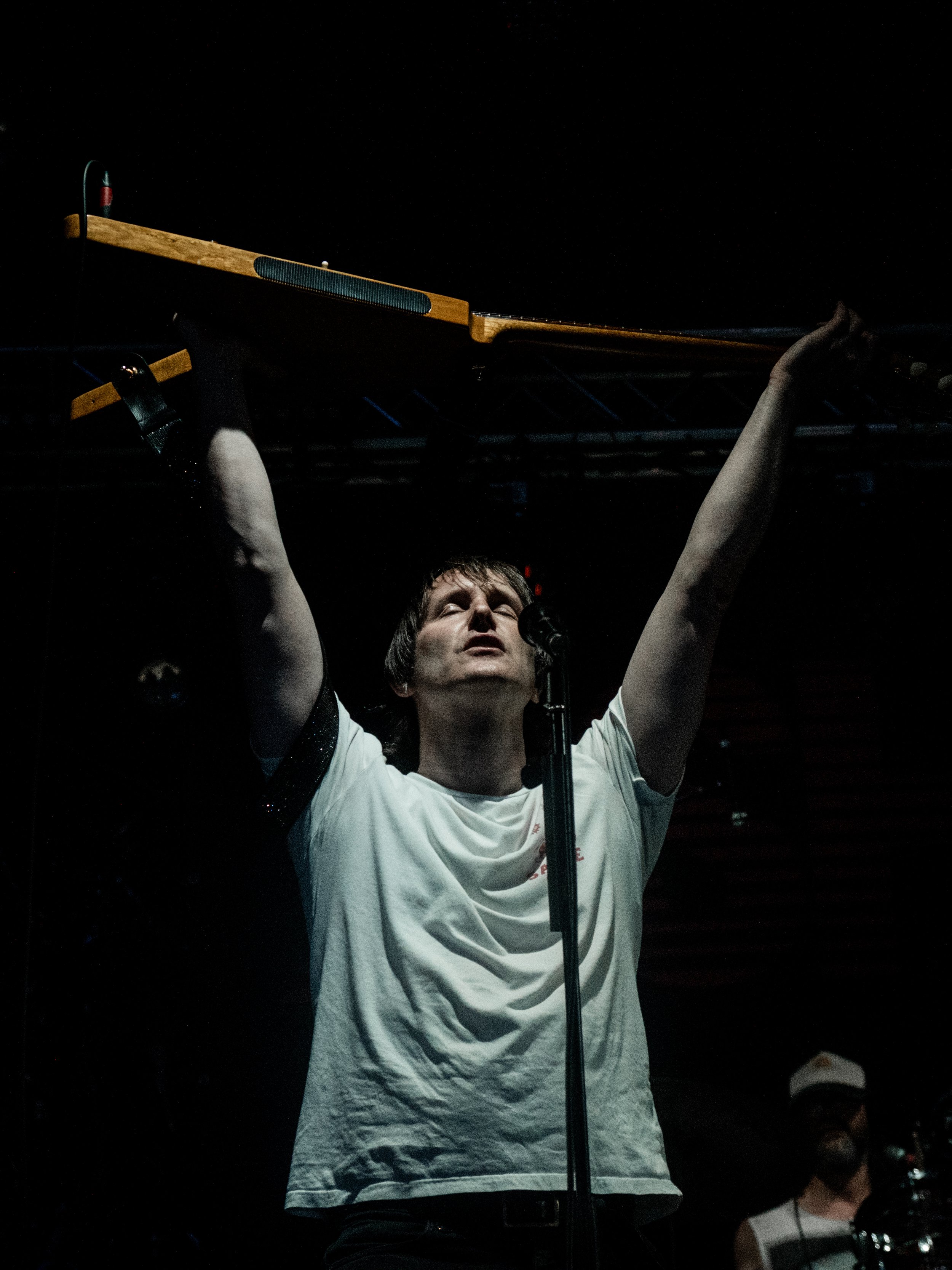 A man on stage holding a long wooden object above his head with both hands, wearing a white t-shirt and standing in front of a microphone.