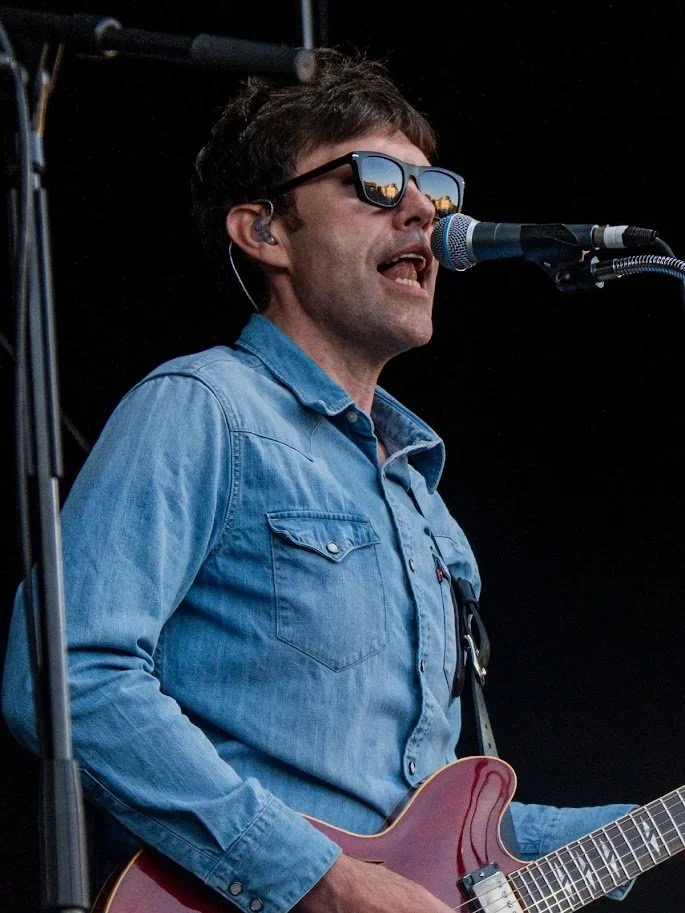 Male musician singing into a microphone, wearing sunglasses, a blue denim shirt, playing an electric guitar on stage.