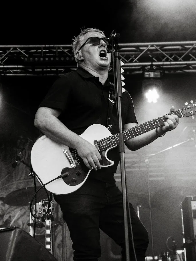 A male musician performing on stage, singing into a microphone and playing an electric guitar. The scene is black and white, with stage lights and equipment in the background.
