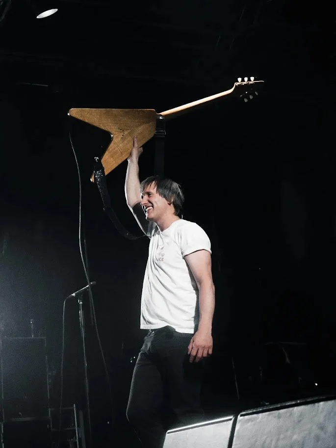 A person holding a guitar headstock up in the air, smiling on a stage with dark background.